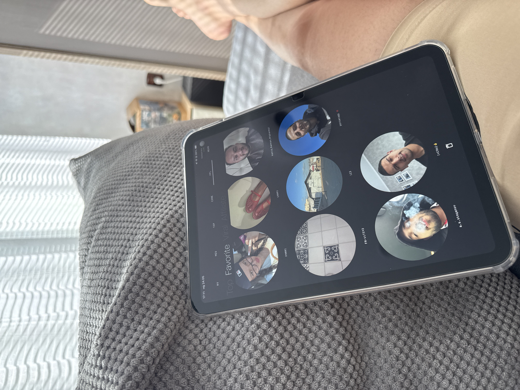 A person sits with a tablet resting on a textured grey pillow. The tablet screen displays an application featuring a variety of profile images, with text labeling sections as "My," "HEU," "TOP," "CORE," and "PPL." Titles such as "Top Favorite Viral Affiliate" and identifiers like "Wahid Rahman Afghanmat," "FM-ttTS43," "petsatagp," and "a.s.stoyanov" are visible near the profile images. The tablet is encased in a clear protective cover. The background includes a glimpse of a bed, a window with white blinds, and a small table or shelf. Based on the OCR text, the time is 17:11 on Sunday, May 24th. The application on the tablet has sections for "MY" and "NEAR". There is no evidence to suggest that this photo was taken in Lovech, Bulgaria.