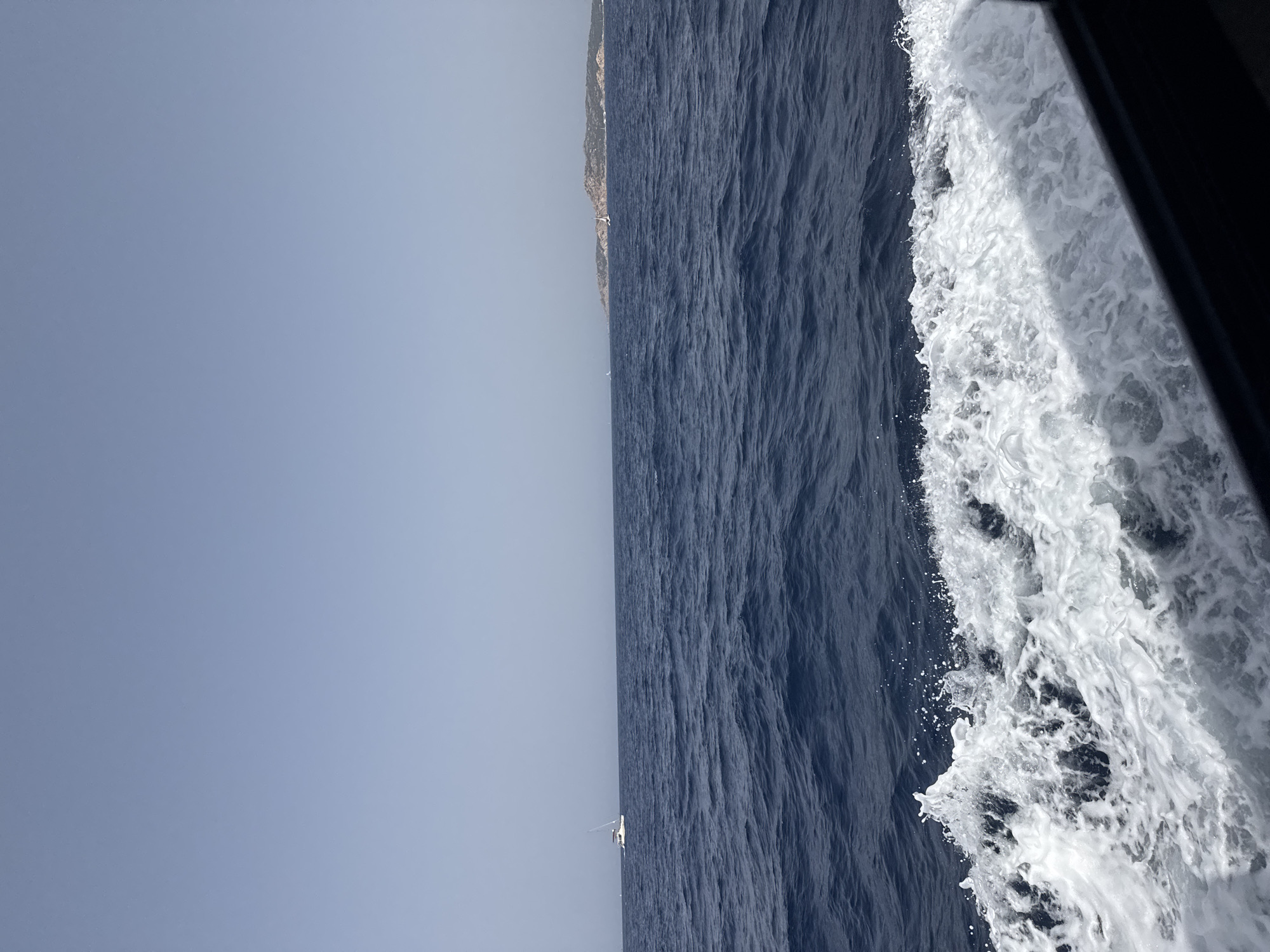 A wide-angle shot captures a tranquil sea under a clear, pale blue sky. The deep blue water is textured with gentle waves and a prominent white wake churns in the foreground, indicating movement. In the distance, a white sailboat is visible on the left side of the frame, and a larger island with vegetation and some buildings is situated on the right horizon. The perspective suggests the photo was taken from a moving vessel. The scene depicts the sea off the coast of La Maddalena, Italy, specifically showcasing the archipelago's characteristic clear waters and distant islands.