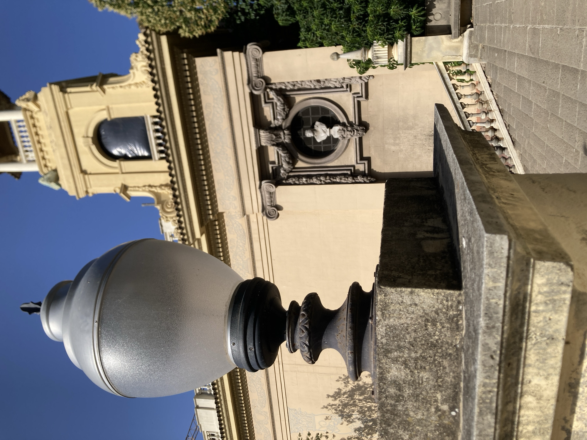 This is a medium-shot, eye-level, outdoor, natural light photograph of a street lamp and a building in Barcelona, Spain. The lamp is in the foreground, on the left side of the frame. It has a frosted glass shade and a dark metal base. The building is in the background, a light yellow color, with a neoclassical architectural style. It has a tower with a clock and arched windows. There is a circular medallion with a bust of a man on the facade of the building. A stone staircase with a decorative railing leads up to the building. The sky is clear and blue. The overall impression is one of historical grandeur and daytime beauty.