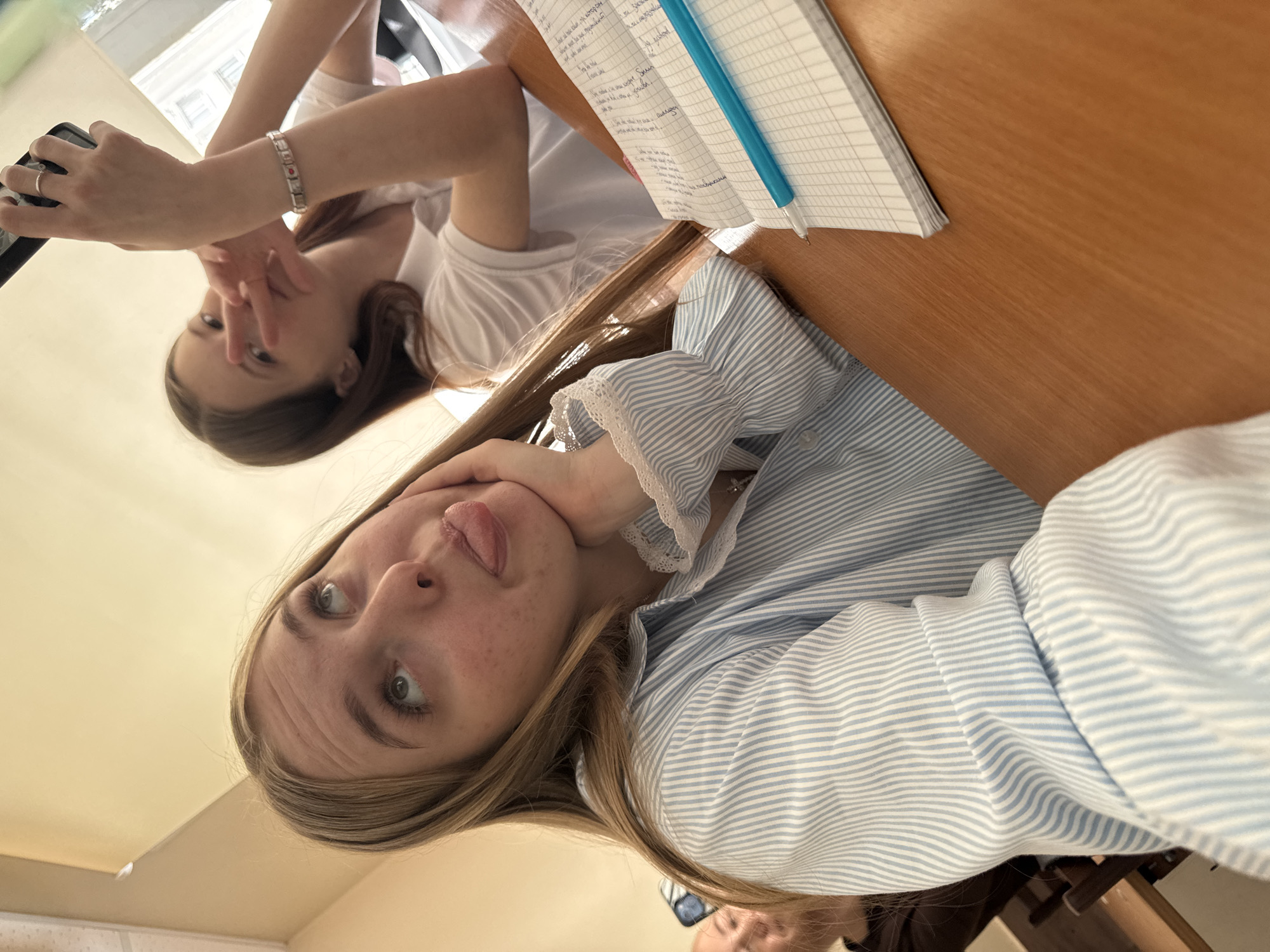 Two women pose for a selfie. One woman with long blonde hair sticks her tongue out and rests her chin on her hand. She wears a striped blue and white shirt with lace trim. Behind her, a second woman with dark hair holds a cell phone and makes a peace sign. They sit at a wooden table with a notebook and blue pen on it. Based on the Cyrillic script in the notebook, the location is likely Russia. The architectural details of the buildings visible through the window suggest the city of Perm.