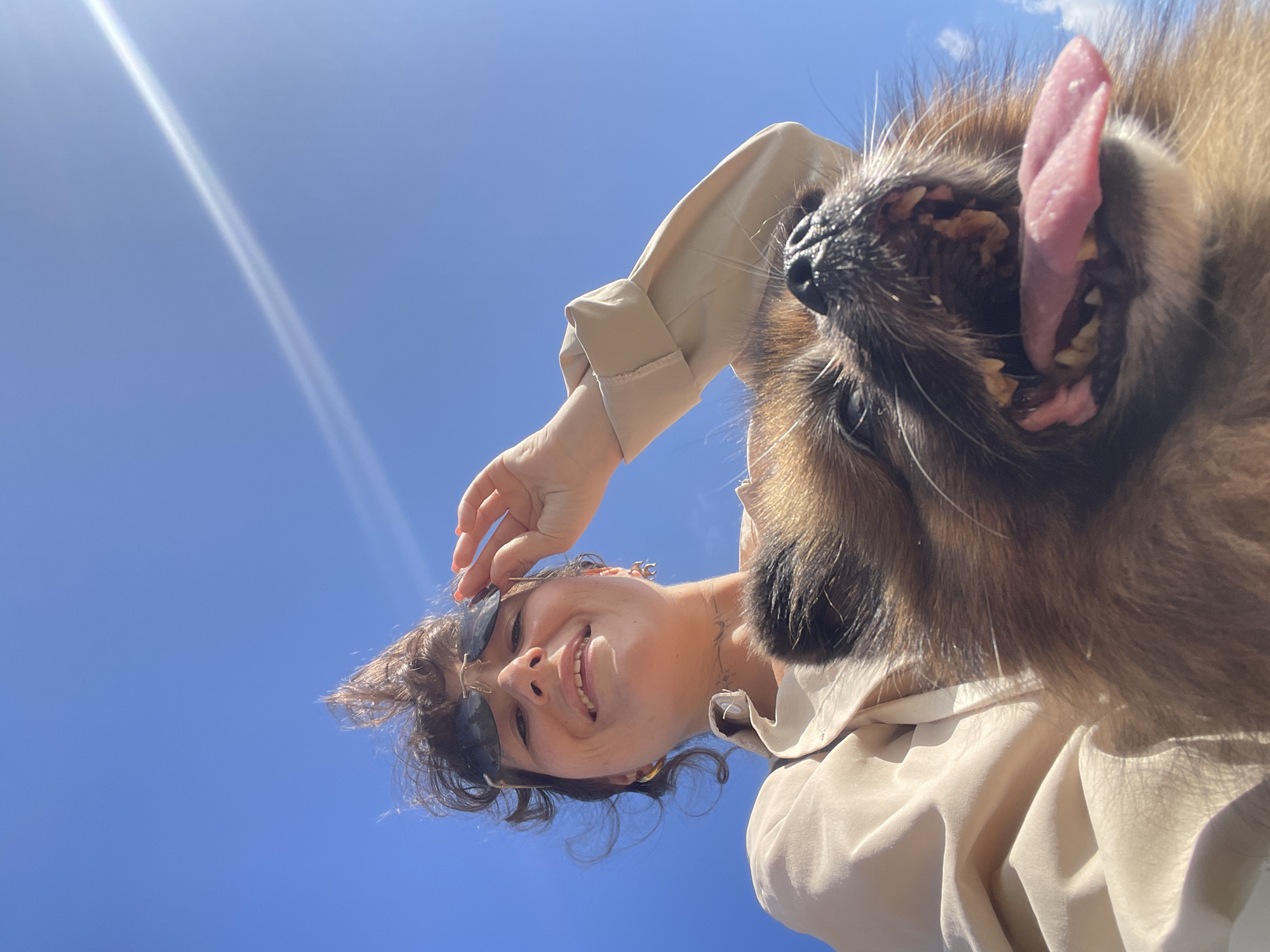 In Rawai, Thailand, under a clear blue sky with wispy clouds, a low-angle shot captures a woman and a dog. The woman, with dark, messy hair and sunglasses perched on her head, smiles up at the camera, her right hand adjusting her sunglasses. She wears a light-colored collared shirt. Beside her, a dog's face fills the foreground, its mouth open, tongue lolling out, and teeth visible. The dog's fur is a mix of brown and black. The overall impression is a candid, sun-drenched moment.