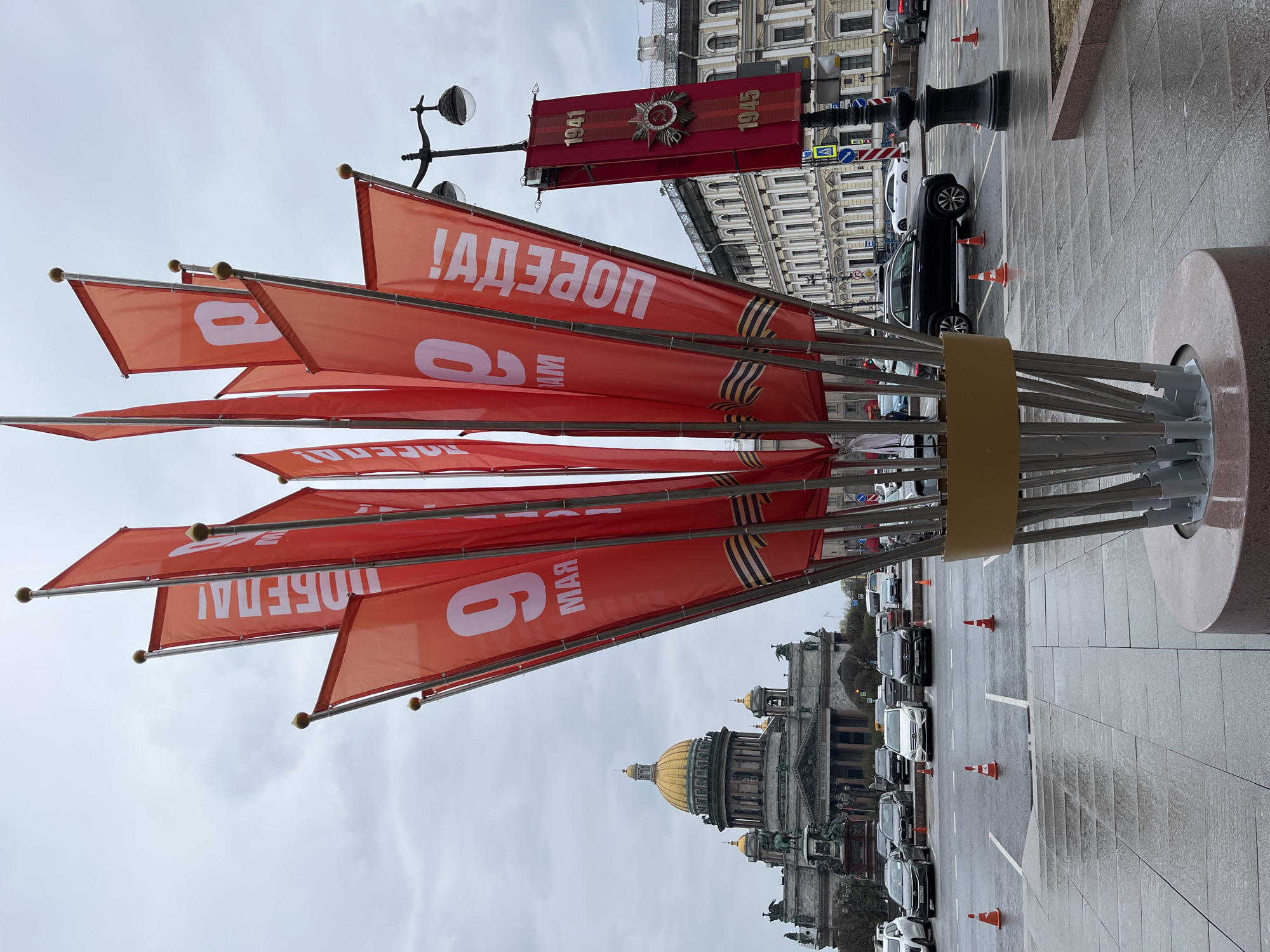The image depicts a street scene in Saint Petersburg, Russia. In the foreground is a display of red flags with white Cyrillic lettering, the number "9," and the words "MAY" and "VICTORY!" Each flag is trimmed with the St. George ribbon. In the background, the golden dome of Saint Isaac's Cathedral is visible. The street is wet, and traffic is visible, including cars and orange traffic cones. A red banner displaying "1941-1945" is affixed to a lamppost. The sky is overcast.