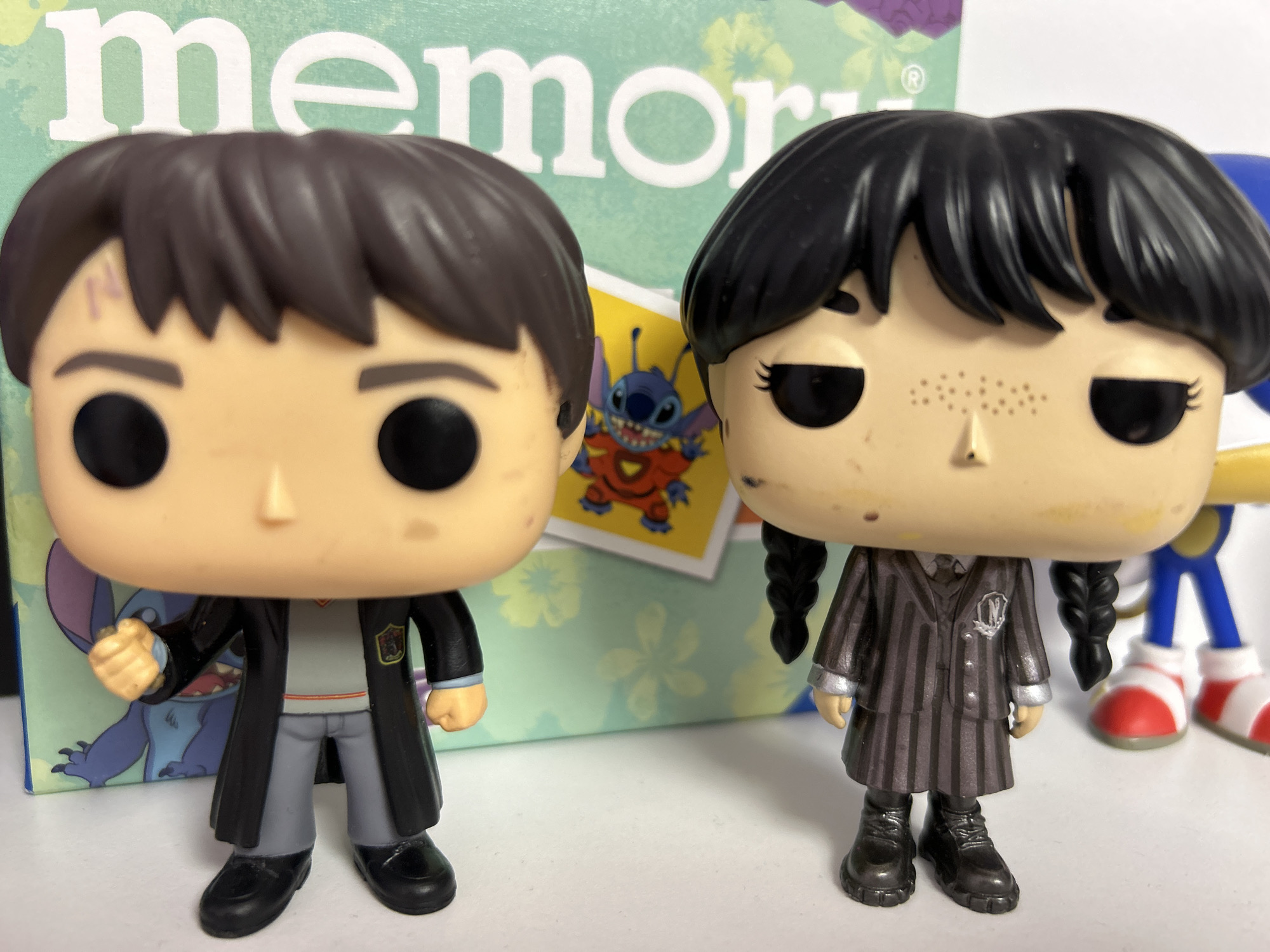 The image shows two Funko Pop figures. The figure on the left is Harry Potter, wearing his Hogwarts uniform and a black robe. He has a lightning bolt scar on his forehead. The figure on the right is Wednesday Addams, wearing her signature black and white striped dress. Behind the figures, a box with the word "memory" is visible, along with a small image of Stitch from Lilo & Stitch. Further in the background, the legs of a Sonic the Hedgehog figure can be seen. The figures are placed on a white surface, likely a table or shelf. The overall scene is well-lit, with the focus on the Funko Pop figures. There is no indication of the city of Burgas, Bulgaria, in the image.