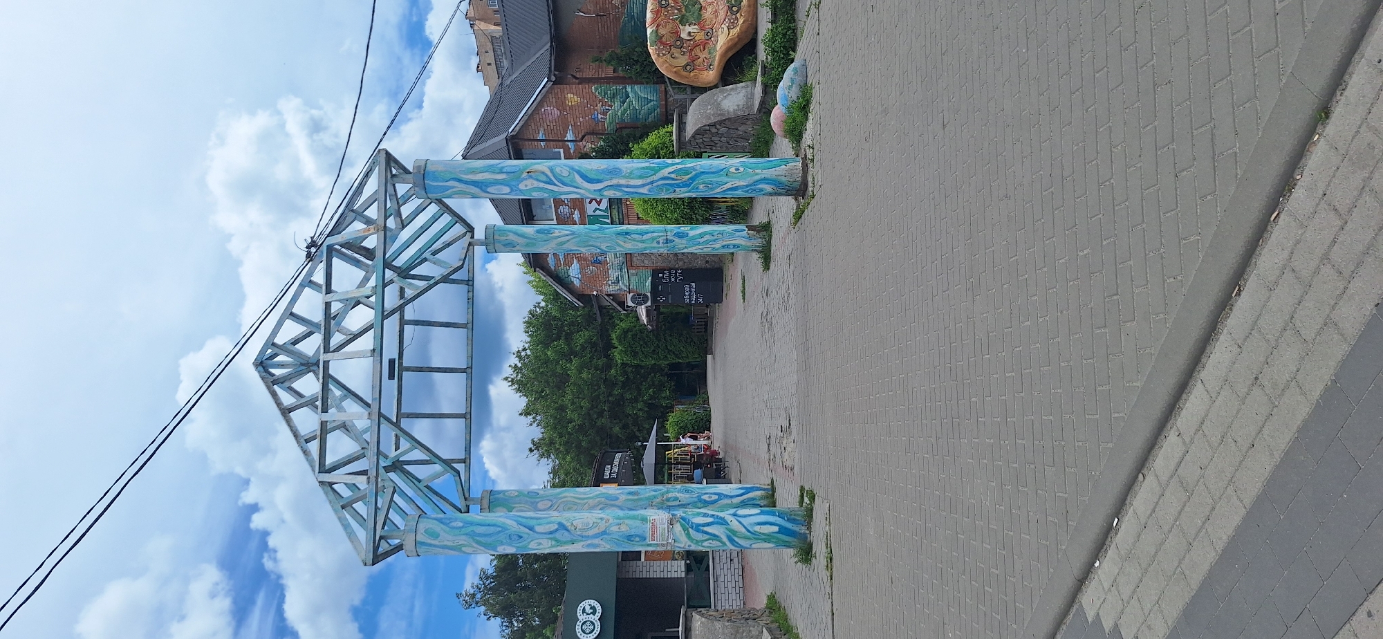 The image depicts an outdoor scene featuring a decorative archway with blue and white painted columns supporting a metal frame structure. The ground in front of the archway is paved with gray brickwork. In the background, there are buildings, trees, and a cloudy sky. One building has a brick facade decorated with a mural. To the right of the archway, there is a large, colorful, mushroom-shaped sculpture. The scene appears to be in a public area, possibly a park or a recreational space. The image may have been taken in Bila Tserkva, Ukraine, given the context. There are no people clearly visible interacting with the archway or sculptures.