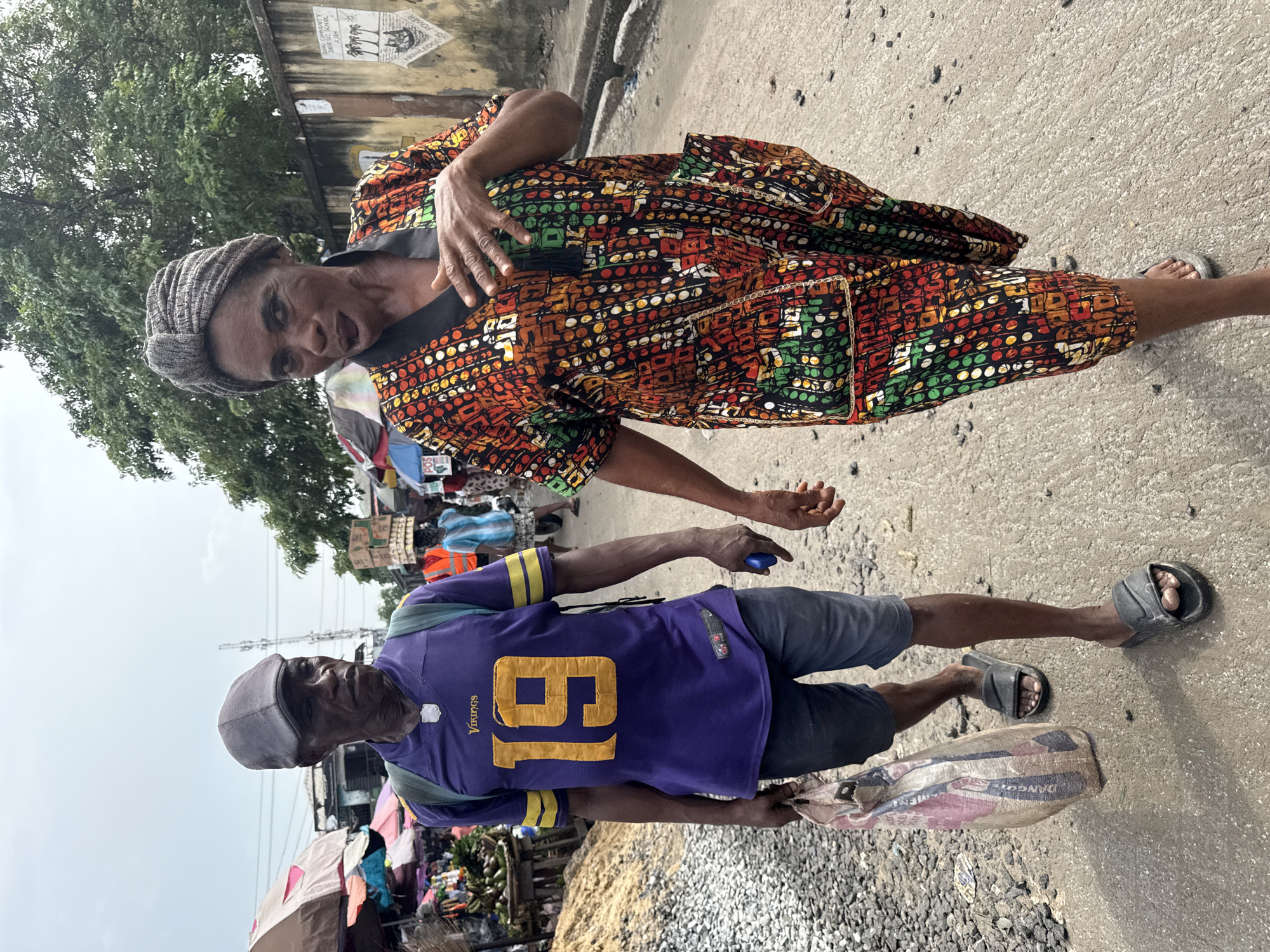 Two individuals, an older woman and an older man, are captured standing on what appears to be a paved street or market area in Ado, Nigeria.
The woman, positioned on the left side of the frame, is facing the camera with a gentle expression. She wears a vibrant, multi-colored tunic or dress featuring geometric and dotted patterns in shades of red, orange, green, and black. Her head is adorned with a textured grey turban-style headwrap. Her right hand is raised towards her chest.
To her right, slightly behind and to the side, stands a man. He wears a purple American football jersey with the number "19" prominently displayed in yellow, and the word "Vikings" is visible on the chest. He is dressed in blue shorts, a grey flat cap, and dark sandals. A small blue object is visible in his left hand, and he carries a patterned sack that clearly reads "DANGOTE CEMENT." He appears to be either walking or standing, with a neutral expression.
The background suggests a bustling outdoor environment. Lush green trees are visible under an overcast sky, indicating daytime. Behind the woman, a wall features several posters, one of which partially displays "HOME COMMUNITY JUNIOR SCHOOL" and "KNOWLEDGE." Further back, various makeshift stalls and market structures can be discerned, consistent with a local street market. The ground is a mix of concrete and scattered small pebbles.
