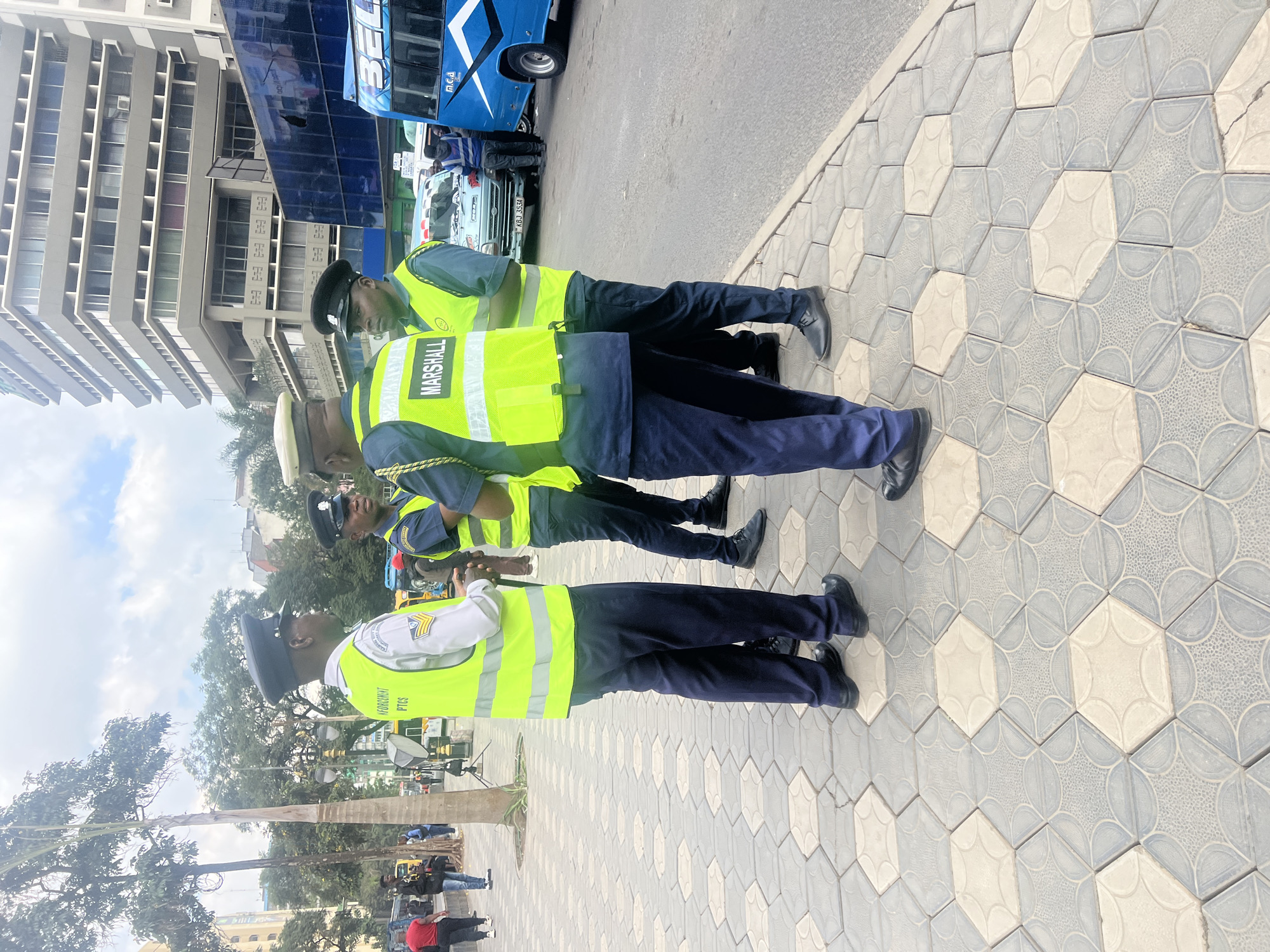 The image shows a row of four uniformed city of Nairobi enforcement officers, possibly traffic marshals, standing on a patterned tiled sidewalk. They wear dark blue uniforms, light-colored caps, and bright yellow vests with the word "MARSHALL" printed on the back of at least one. The officers appear to be engaged in a discussion or observation, with at least one holding a mobile device. Behind them, there is a street with parked vehicles and a blue bus, as well as a tall building. The environment suggests an urban setting, likely within the city of Nairobi.