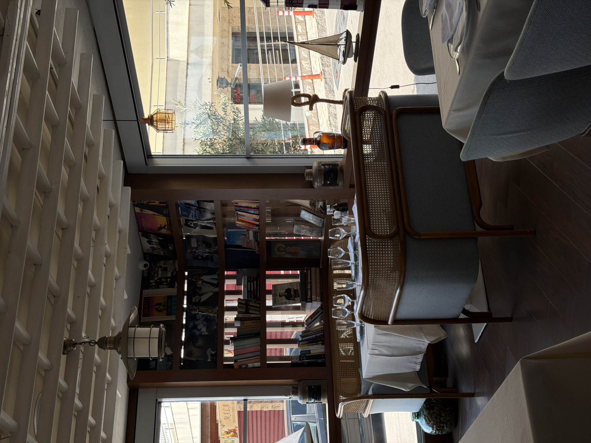 The image shows the interior of a restaurant, likely in Rafailovići, Montenegro, given the location tag. The restaurant features a floor-to-ceiling bookshelf filled with books and various decorative objects. A window runs alongside the bookshelf, offering a view of the street outside with its signage and buildings.
The scene includes dining furniture, such as tables with white tablecloths and chairs with rattan detailing. Wine glasses are arranged on the table. A decorative lamp with a white shade and a rope base sits on the bookshelf near the window. Another lamp fixture hangs from the ceiling.
The overall ambiance suggests a sophisticated and cozy dining environment. The image is taken from an elevated perspective.