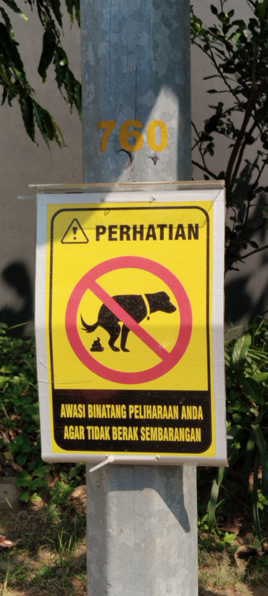A yellow sign with a black border is attached to a galvanized metal pole. The sign has the text "PERHATIAN" (Attention) at the top, with a black triangle and exclamation mark to its left. Below this, there is a red circle with a diagonal line through it, superimposed over a black silhouette of a dog defecating. The text at the bottom of the sign reads "AWASI BINATANG PELIHARAAN ANDA AGAR TIDAK BERAK SEMBARANGAN" (Watch your pet so it does not defecate carelessly). The pole has the number "760" painted in yellow on its upper section. The scene appears to be outdoors, with greenery and foliage visible in the background, suggesting a park or public area. The lighting indicates it is daytime, with shadows cast on the pole. The overall impression is a public service announcement aimed at pet owners. The image was taken in Semarang, Indonesia.