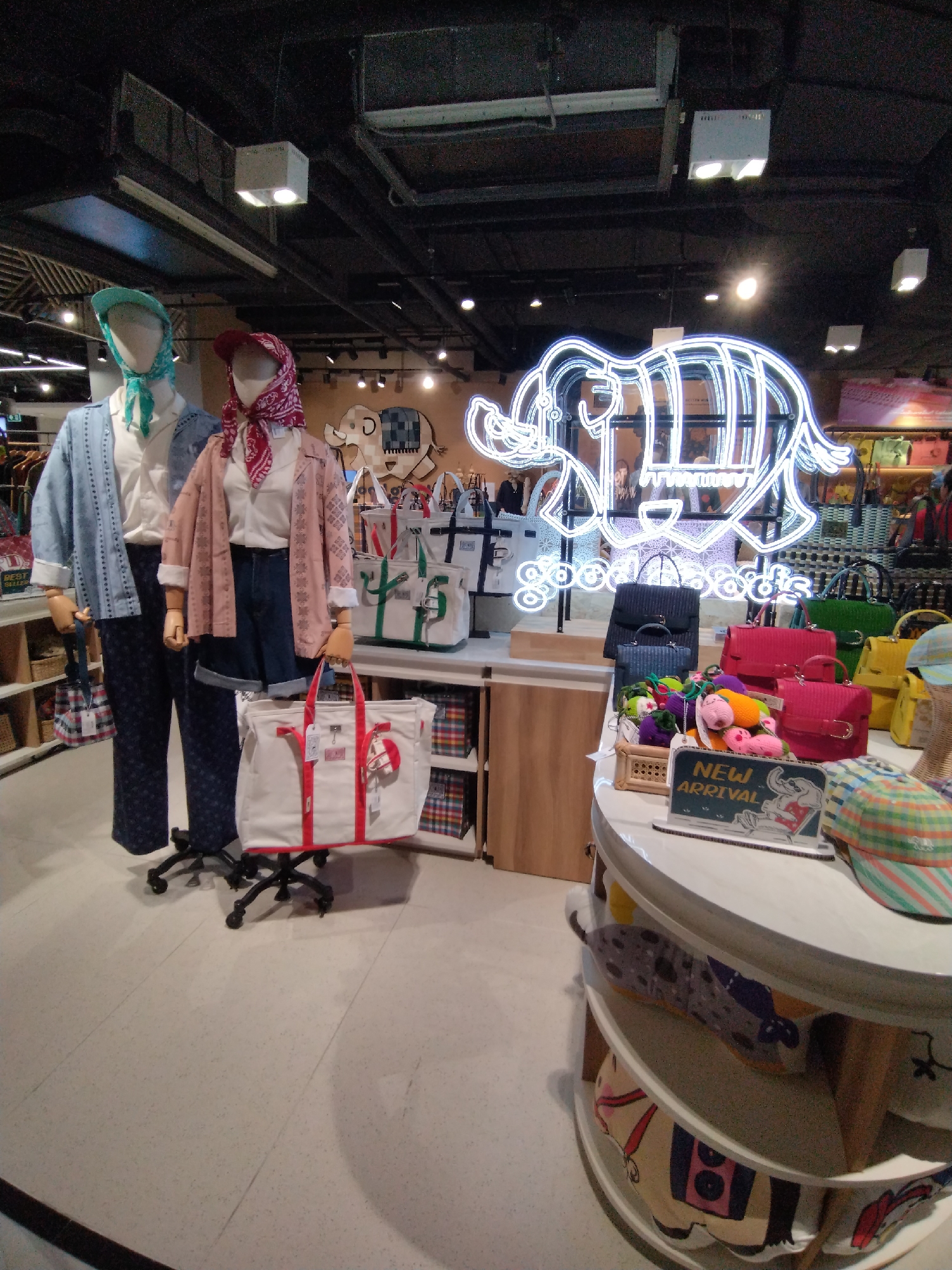 The image depicts the interior of a retail store, likely a boutique or department store, exhibiting clothing and accessories. Prominently featured are two mannequins dressed in casual apparel. The mannequin on the left wears a light blue patterned jacket over a white shirt and dark patterned trousers, accessorized with a teal headscarf. The mannequin on the right is dressed in a pink patterned jacket, a white shirt, and denim shorts, with a red headscarf.
In the background, a large neon sign in the shape of an elephant with the words "good goods" illuminated in blue light is visible. Various handbags, predominantly in canvas and bright colors like red, green, and yellow, are displayed on shelves and a round tiered display table. A "NEW ARRIVAL" sign is placed on the display table, accompanied by small stuffed toys.
The environment appears to be well-lit with overhead lighting fixtures. The floor is tiled with a light-colored speckled pattern. Shelving units stocked with items are visible along the walls. The overall impression is that of a modern, well-organized retail space showcasing trendy merchandise.
The scene is consistent with the type of retail outlets found in tourist areas such as Patong, Thailand, which are known for their vibrant shopping experiences catering to both locals and international visitors. While the image itself does not provide definitive location markers, the style of the store and the merchandise are typical of what one might find in such a location.