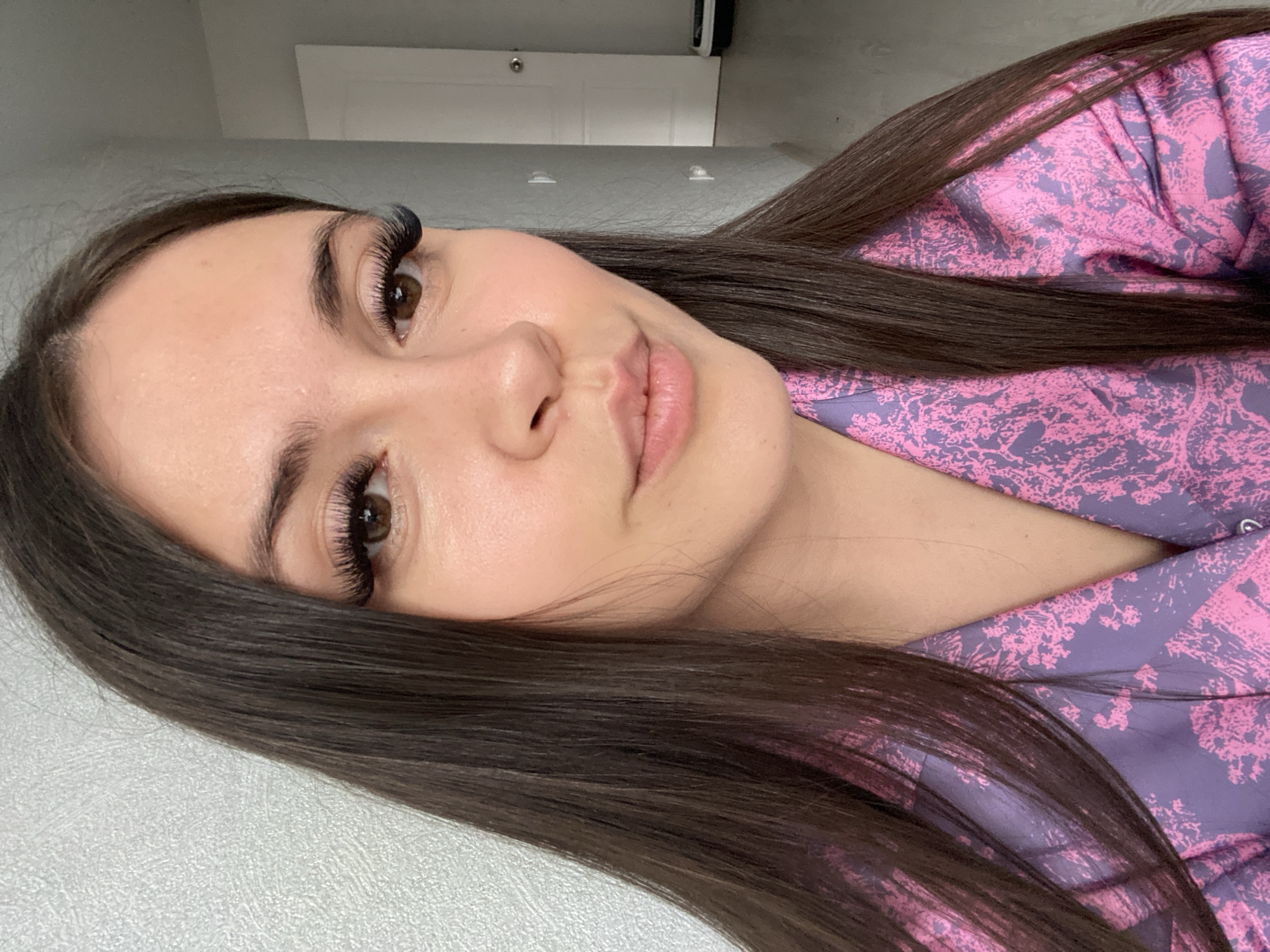 A close-up, slightly angled portrait depicts a young woman with fair skin, brown eyes, and dark brown hair. She is wearing eyelash extensions. Her expression is neutral with a slight smile. She wears a pink and purple patterned garment. The background includes a light gray or white surface and a partially visible white door in the distance. There is no information available in the image to confirm the location within Chaikovsky, Russia.