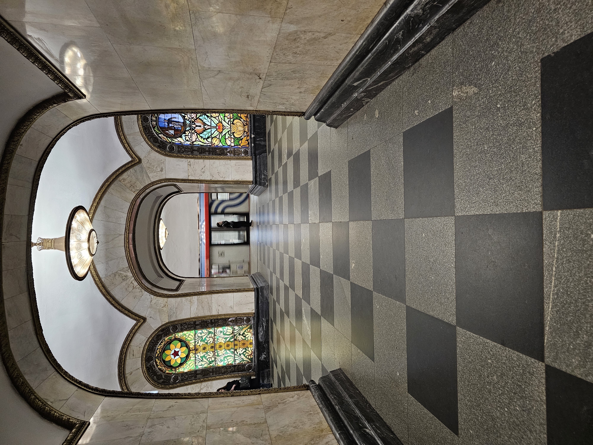 The image shows an interior view of Komsomolskaya station in Moscow, Russia. The perspective is vertically oriented, looking down a hallway or corridor. The hallway features black and gray checkered flooring. On the left side, there are ornate, arched doorways with stained glass windows. The walls are marbled and decorated with gold trim. A chandelier hangs from the ceiling. At the end of the hallway, there are figures of people.