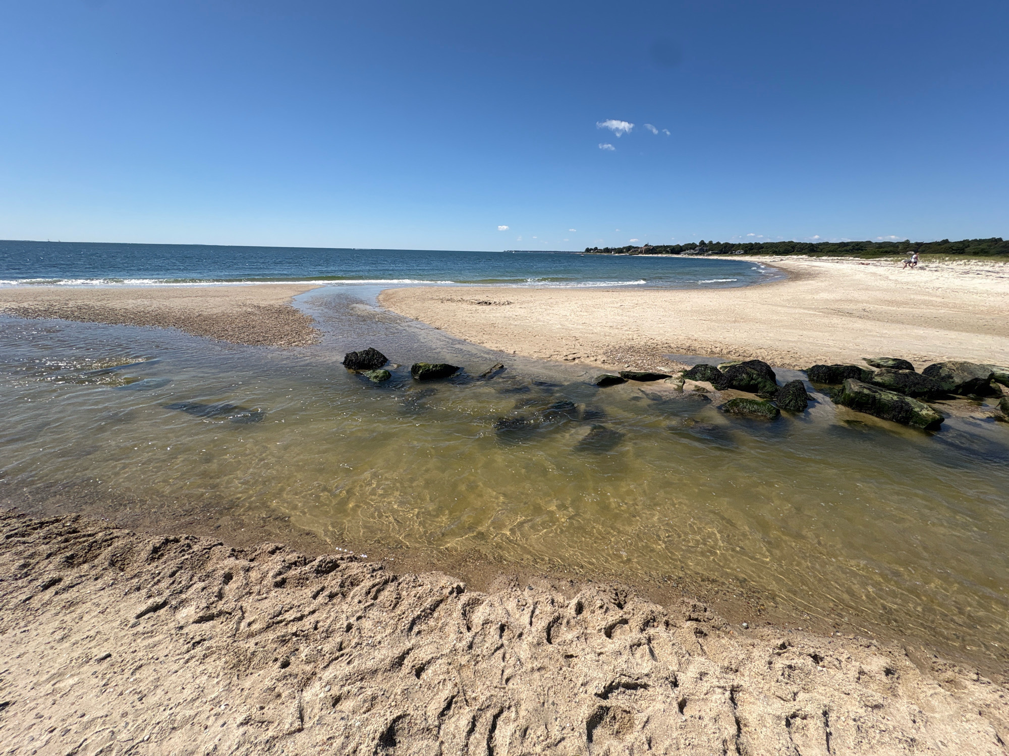 The image depicts a scenic view of Ocean Beach Park in Waterford, Connecticut, on a clear, sunny day. In the foreground, a sandy beach is evident, with textured sand showing signs of footprints or tire tracks. A shallow stream of water, with a greenish-brown hue, flows from the left towards the ocean, bordered by dark, moss-covered rocks on its right bank. The water in the stream is clear enough to reveal the sandy bottom and some submerged rocks.
Beyond the stream, a wider expanse of sandy beach stretches towards the horizon. The ocean, a deep blue, meets the shore with gentle waves. In the distance, along the coastline, a line of trees forms a lush green border, with some buildings visible nestled among them, suggesting a developed area beyond the natural landscape. The sky is a vibrant, cloudless blue, with a single, small white cloud visible in the upper right portion of the frame. Two people are faintly visible on the beach in the far distance, near the tree line, suggesting recreational activity. The overall scene conveys a peaceful and serene atmosphere of a summer day at the beach.