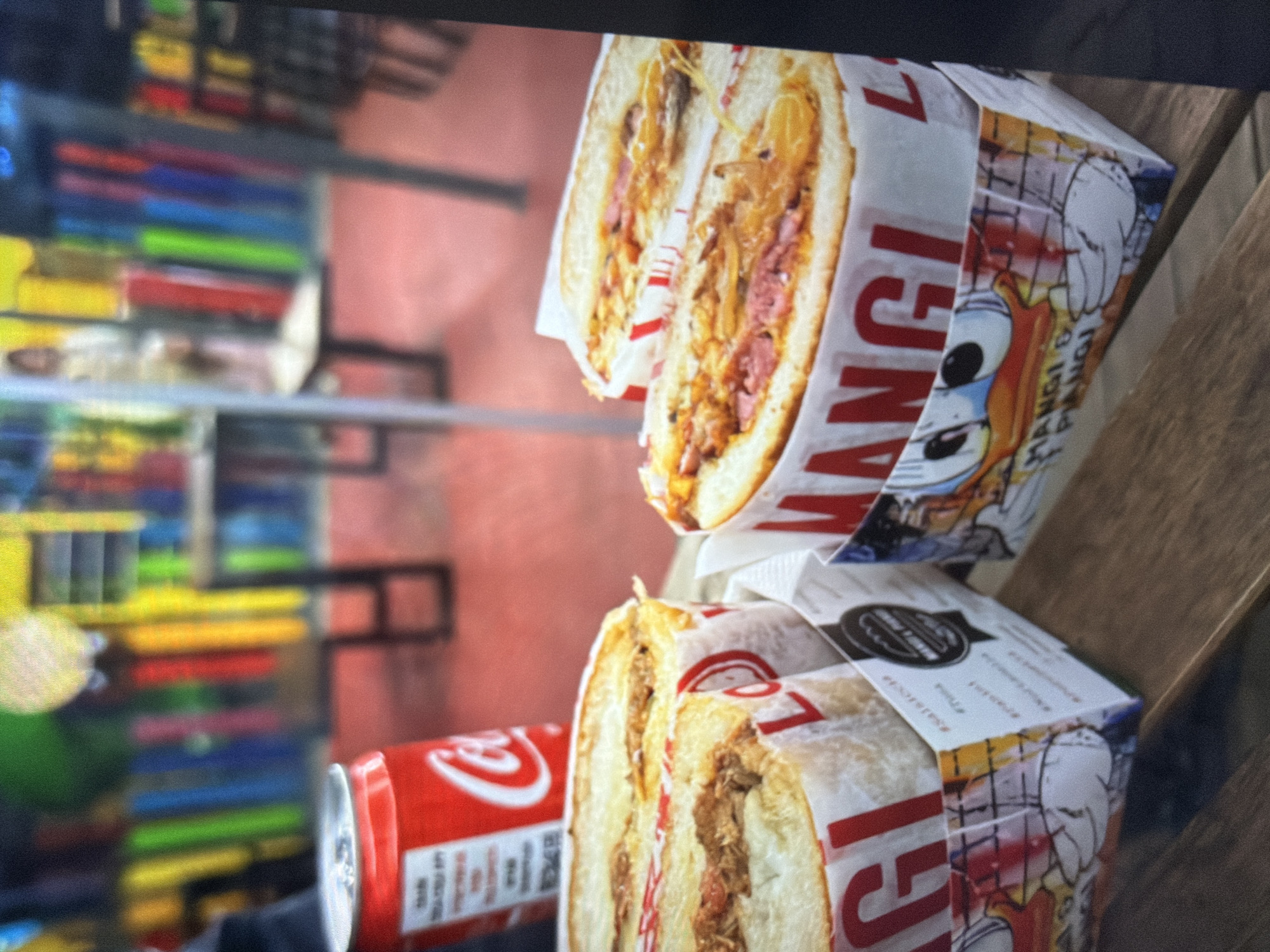 The image shows two halves of two sandwiches in branded packaging stacked on top of each other. Next to the bottom sandwich is a can of Coca-Cola. In the blurred background is a vending machine containing several rows of items. The sandwich packaging reads "Mangi e Piangi" which translates to "Eat and Weep." The location of the city is unknown.