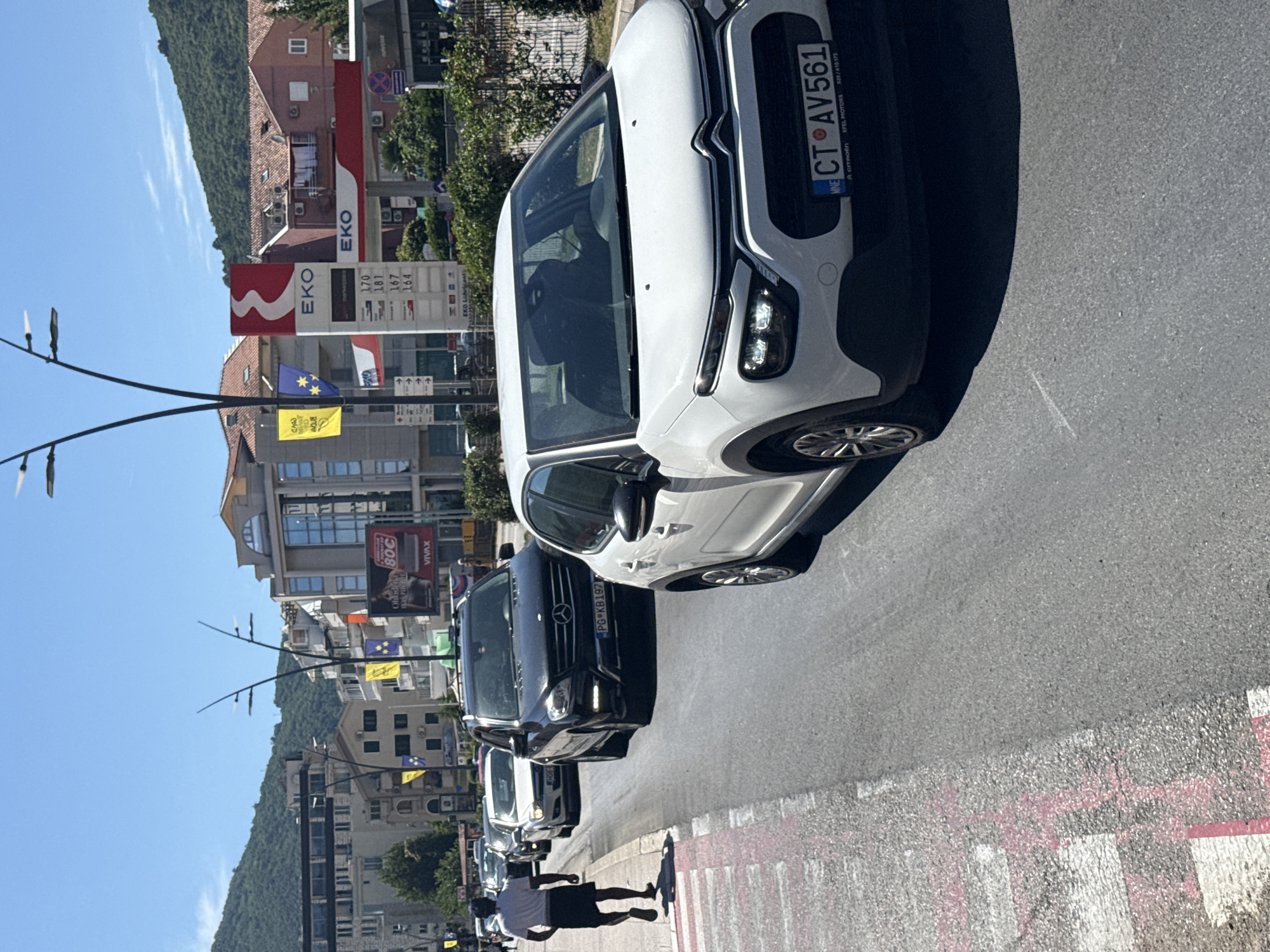 In Budva, Montenegro, on a sunny day, traffic is backed up on a city street. A white Citroen C3, license plate CT AV561, is prominently in the foreground, facing away from the viewer. To its left, a black Mercedes-Benz M-Class, license plate PG KB197, is also stopped in traffic. A light-colored car follows the Mercedes. Further back, several other vehicles are visible, forming a line. On the sidewalk to the left, a person wearing a white t-shirt and black shorts walks away from the camera. Buildings line the street, including a multi-story structure with balconies and a gas station with EKO branding and fuel prices displayed. Flags are visible on poles, including yellow and blue flags. The road appears to be asphalt, with a pedestrian crossing marked with white and pink lines on the left side. Green foliage and hills form the backdrop.
