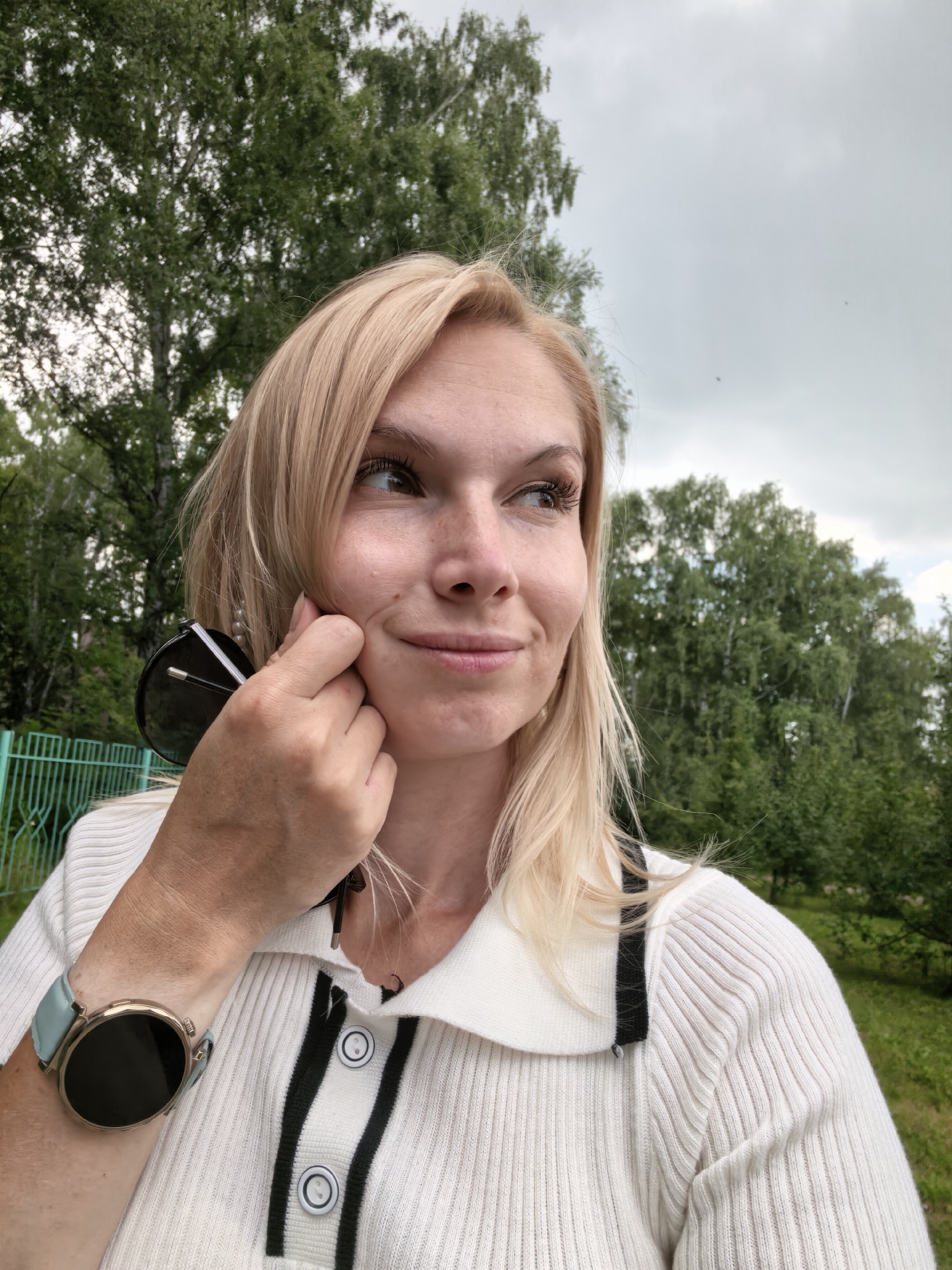 A blonde woman with freckles is taking a selfie outdoors. She is wearing a white ribbed sweater with black trim and buttons. A smart watch with a light blue band is on her left wrist. She is holding a pair of sunglasses up to her cheek with her right hand. The background consists of green trees and a cloudy sky. A portion of a green fence is visible on the left side of the image. The location is Sterlitamak, Russia.