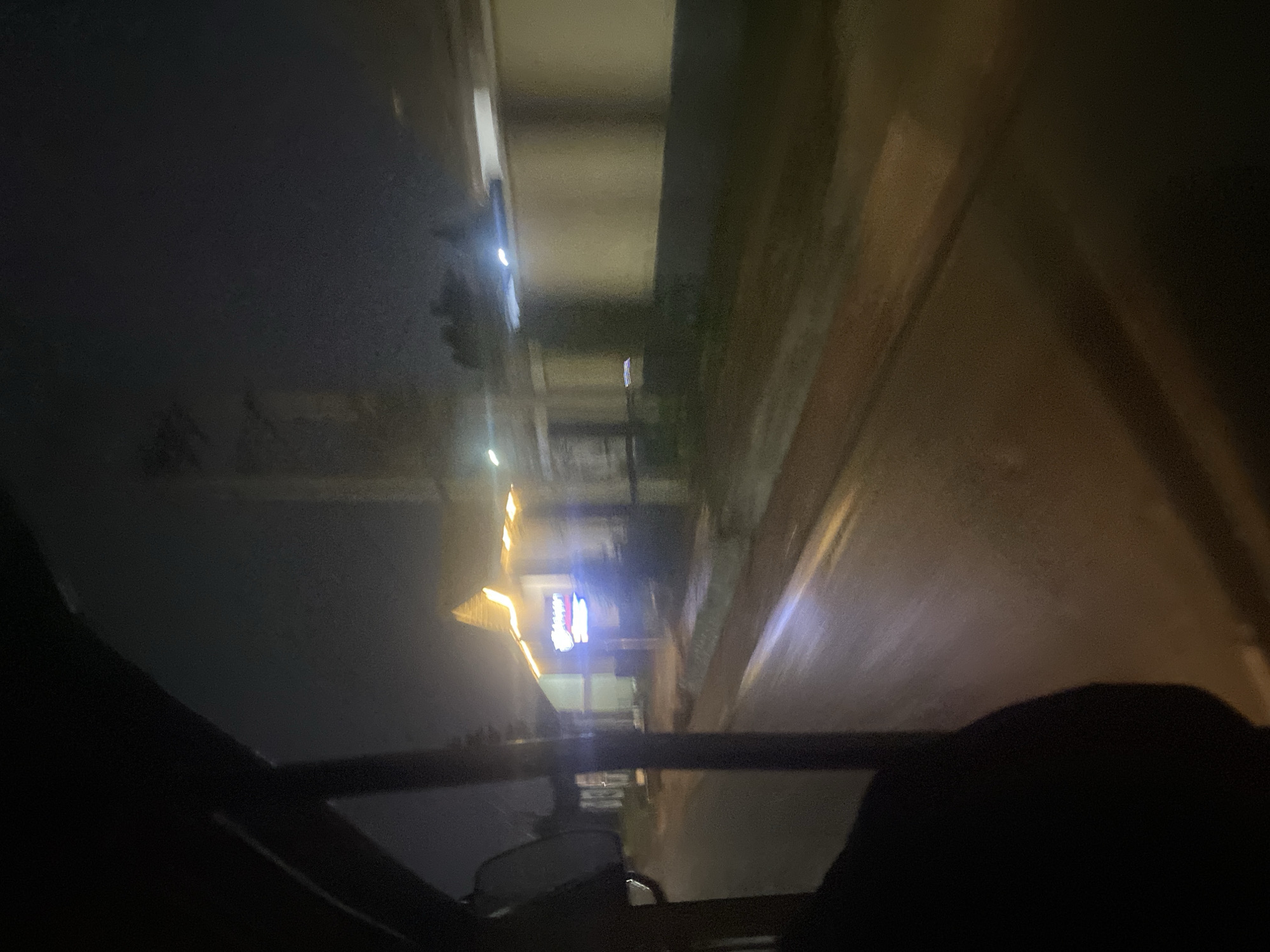 The image depicts a view from inside a vehicle at night, likely in Gura Top, Nigeria. The scene is dimly lit, with artificial lights illuminating a building with a distinctive peaked roof. A neon sign, appearing to be in blue and possibly pink or red lettering, is visible on the building's facade, though the text is not clearly legible. The street in front of the building is wet, reflecting the lights, suggesting recent rain or a damp surface. The surrounding environment appears urban, with hints of other structures and streetlights in the background. The perspective suggests the vehicle is in motion, contributing to the slight blurriness of the scene, especially in the distance. The foreground shows a dark interior of the vehicle, including a portion of the door frame and a side mirror. The overall atmosphere is nocturnal and slightly hazy.