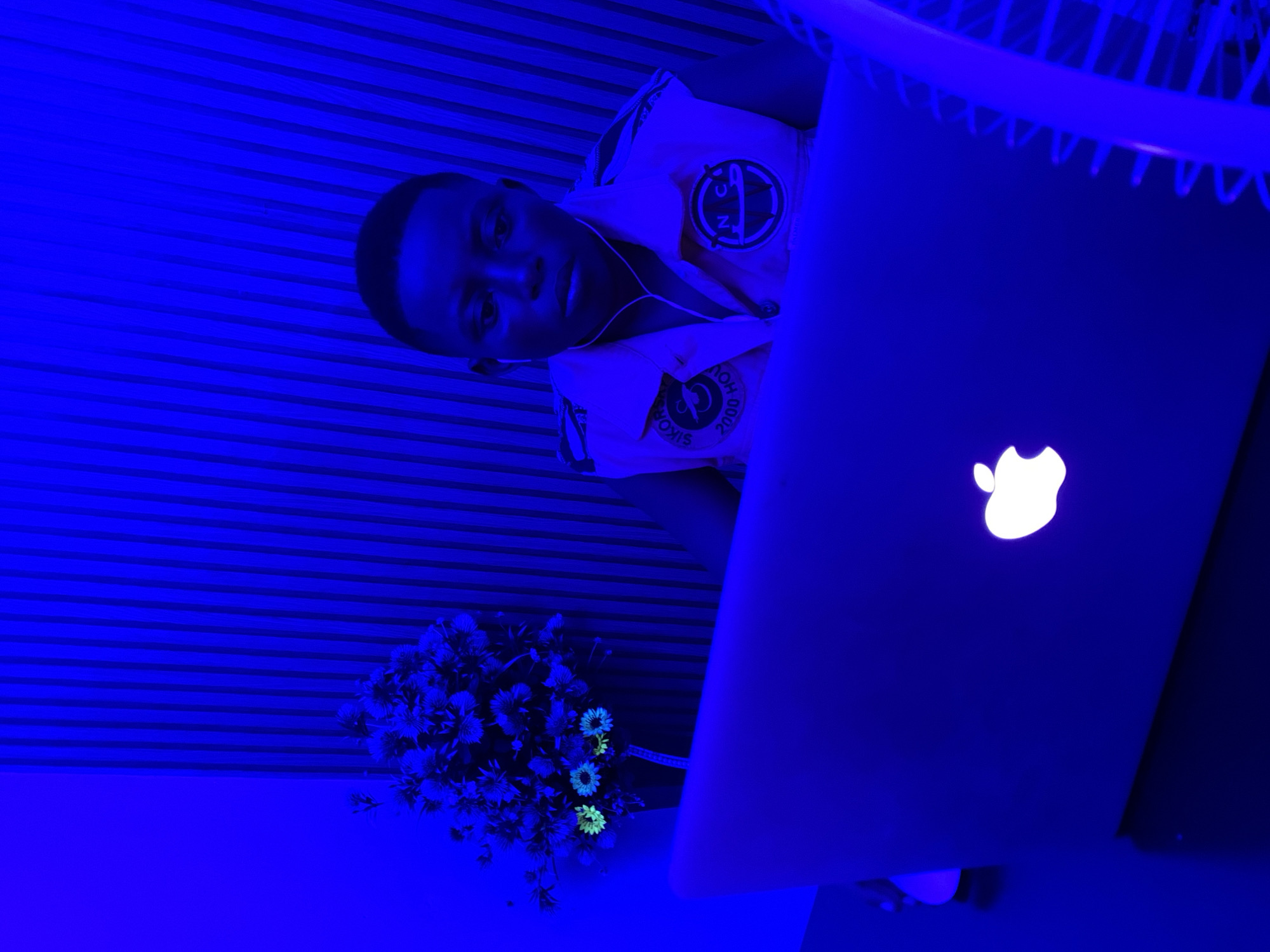 A person is seated at a table using a laptop. The Apple logo on the laptop is illuminated. A bouquet of flowers is on the table beside the laptop. The room is lit with blue light. The location is implied to be in Ikorodu, Nigeria, though the image itself does not confirm this. The person is wearing a collared shirt with patches. The person appears to be wearing earphones. The background has horizontal lines.