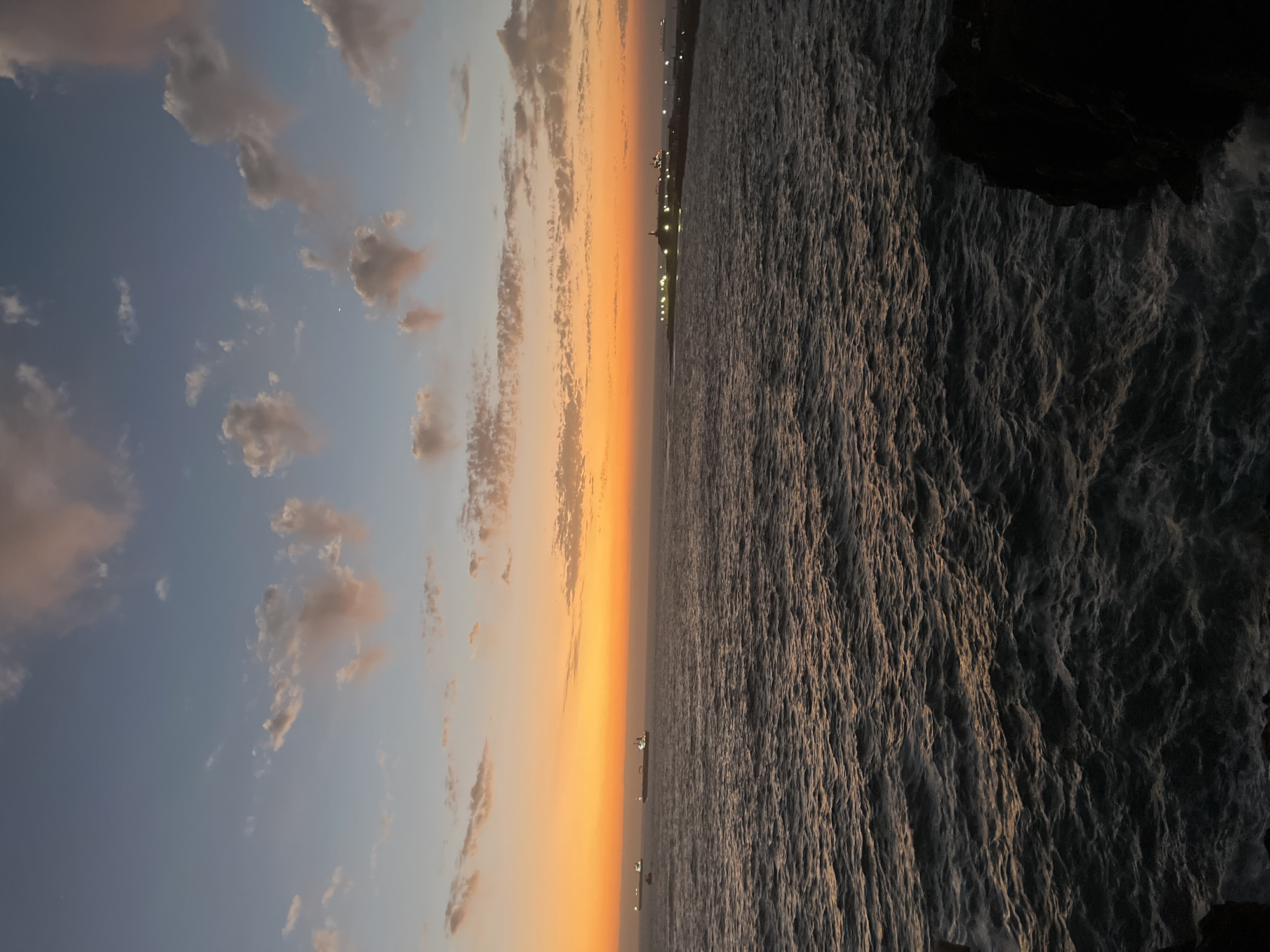 The image shows a sunset scene in Arica, Chile. The sky is divided into two sections: the left side displays a blue sky with scattered white and pink-tinged clouds, while the right transitions to a vibrant orange hue. A body of water, likely the Pacific Ocean, fills the right half of the image with visible waves and reflections of the sunset. In the distance, lights from the city outline the coastline. A dark, rocky formation is visible in the lower right corner, suggesting a cliff or rocky shore. A ship can be seen silhouetted against the sunset in the distance.