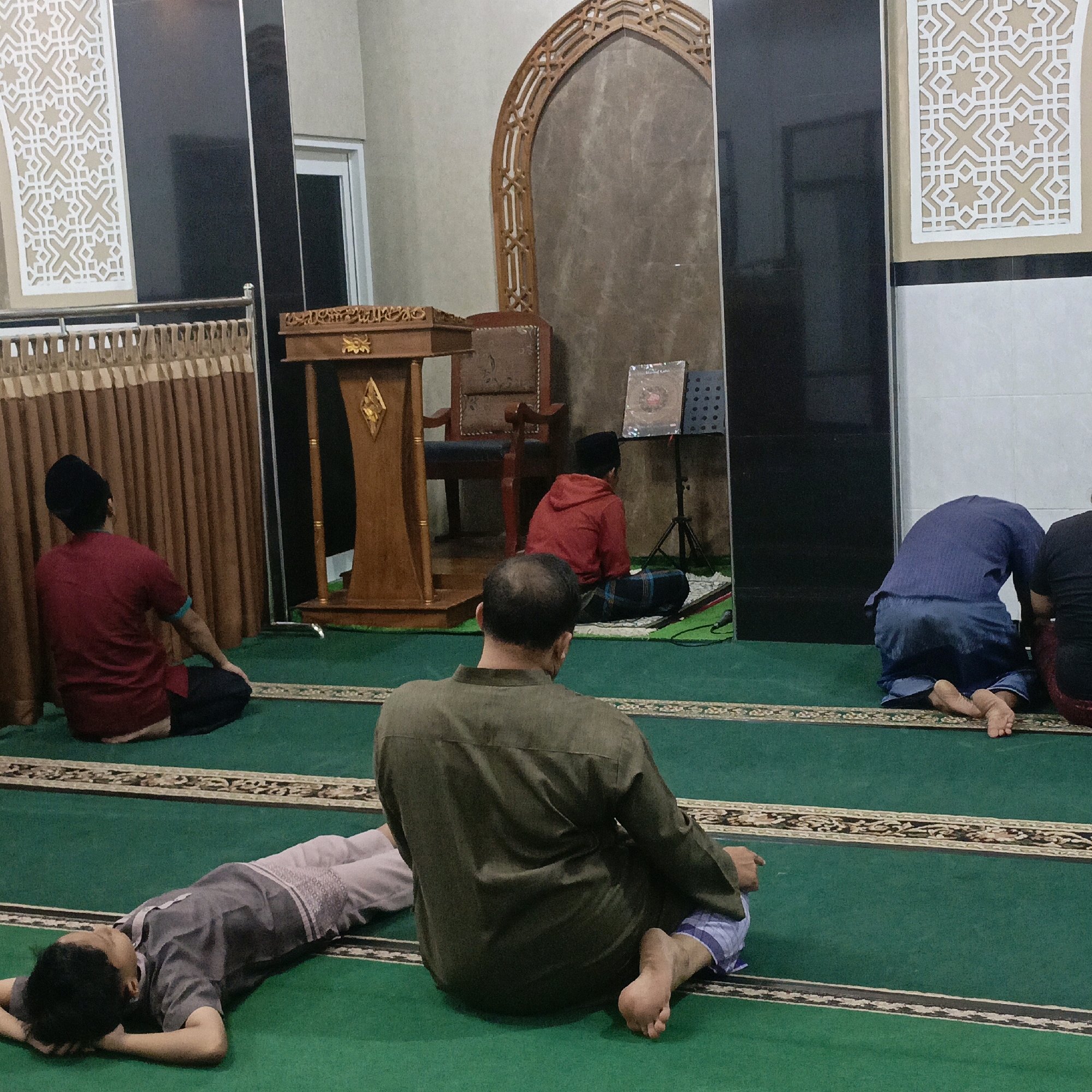 The image depicts an indoor scene, likely a mosque, with multiple individuals engaged in prayer. The floor is covered with a green carpet marked with decorative borders. In the foreground, a person in an olive-green shirt and checkered pants is seated, facing away from the camera. To their left and slightly further back, a person in a grey shirt and white pants is lying on their back with hands behind their head. In the middle ground, a person in a red hooded shirt and sarong is seated on a prayer mat, facing a small lectern that holds an open book. To the left, a person in a maroon shirt and dark pants is seated, facing the wall with their back to the camera. In the background, a wooden pulpit stands against a patterned wall, and ornate geometric carvings adorn the walls. To the right, two individuals are visible, one in a dark blue shirt and pants, kneeling, and another in a black shirt, partially visible. The lighting suggests an interior setting, possibly during prayer time. The overall atmosphere is one of prayer and contemplation. This scene could be located in Salatiga, Indonesia, a city known for its significant Muslim population and numerous mosques.
