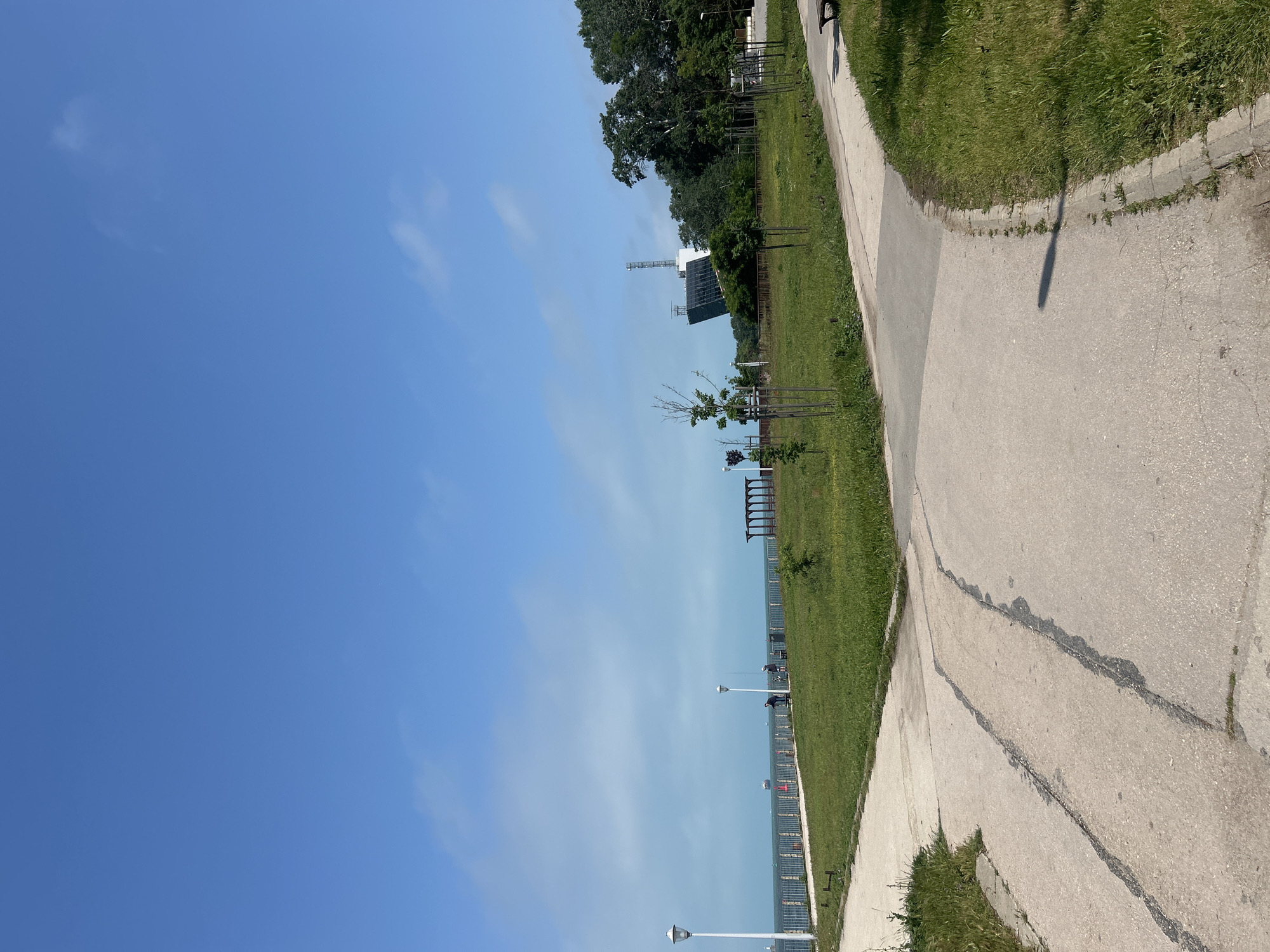 The image displays an outdoor scene, likely a coastal park or promenade, under a bright, clear blue sky with a few faint, wispy clouds. A paved pathway, appearing to be asphalt or concrete, runs along the right side of the frame, showing some signs of wear, including visible cracks and repairs. Adjacent to this path, to the left, is a broad stretch of vibrant green grass.
Beyond the grassy area, a calm body of water, likely the Black Sea given the Varna, Bulgaria context, extends towards the horizon. Along the boundary between the grass and the water, modern street lamps with white caps are intermittently placed, and a fence or low barrier is visible. Further into the grassy expanse, some dark, bench-like structures are present.
In the upper-left background, a dense line of mature green trees forms a treeline. Below this treeline and above the distant water, a prominent white building with a large, dark, possibly reflective or panelled facade stands out. Next to this building is a tall, slender lattice tower, suggesting a potential communication facility, radar station, or observatory. Several smaller, newly planted trees are scattered across the grassy area closer to the foreground.
Long shadows stretch across the path and grass, indicative of either early morning or late afternoon sunlight. A few extremely small, indistinct figures can be faintly seen in the far distance near the water, possibly individuals strolling. No visible text is present in the image. The scene conveys a sense of peaceful outdoor activity and open space.