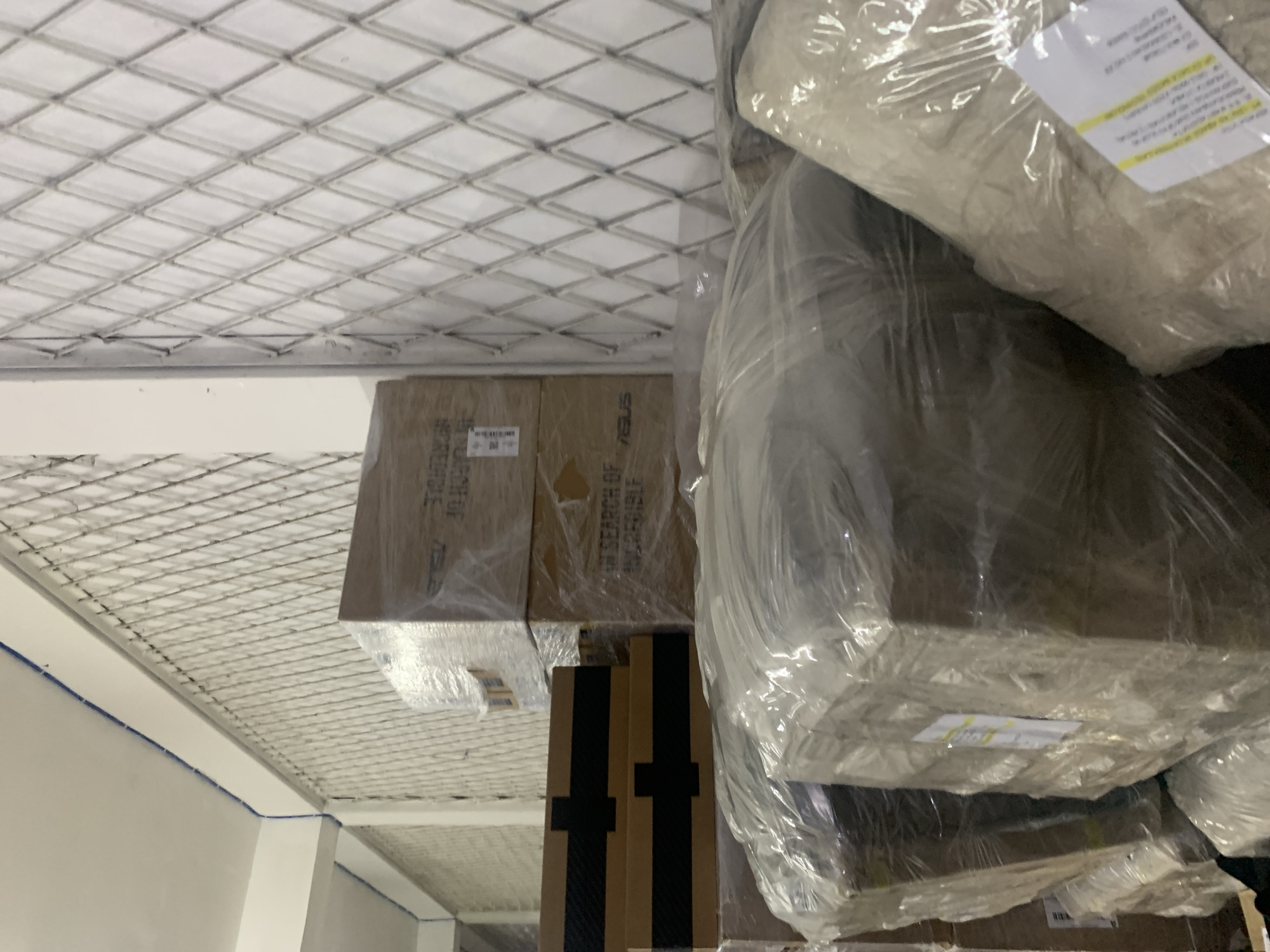 In a dimly lit warehouse, stacks of cardboard boxes are neatly arranged and wrapped in transparent plastic. The boxes vary in size, with some showing labels and markings. The walls of the warehouse are white, with a diamond-patterned metal grate covering a portion of the wall, likely for ventilation or security. Blue electrical conduits are visible along the ceiling and upper wall. The scene appears to be a storage facility or distribution center within the city of Palembang, Indonesia, awaiting shipment or processing.