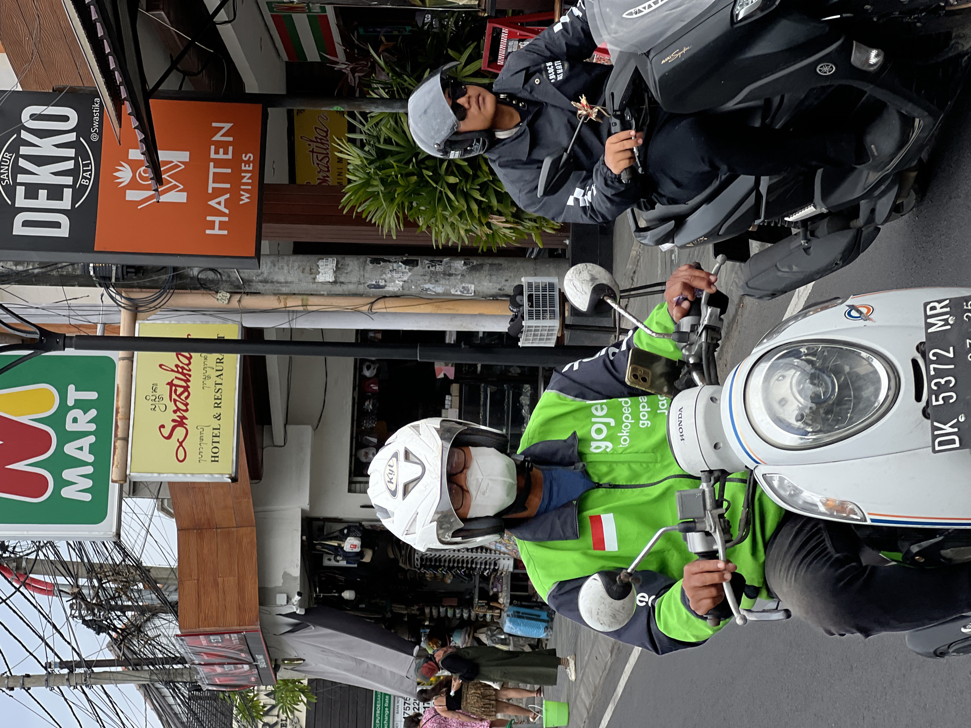The image captures a street scene in Denpasar, Indonesia, likely a commercial area given the signage. Two individuals are riding motorcycles. The foreground features a person wearing a green Gojek uniform, a white helmet with a face mask, and black pants. They are riding a white Honda scooter. To the right, another person is riding a black motorcycle, wearing a black jacket, black pants, a grey helmet, and sunglasses.
Several signs are visible in the background. One prominent sign is for "MART" in green and white. Another sign reads "Swastika HOTEL & RESTAURANT". Further to the right, there is a sign for "DEKKO BALI @Swastika" and below it, a sign for "HATTEN WINES".
The environment appears to be a typical urban street with overhead electrical wires and buildings lining the road. The lighting suggests it is daytime, with slightly overcast conditions. Other people and elements of street commerce, like shops with displays, are visible in the background.