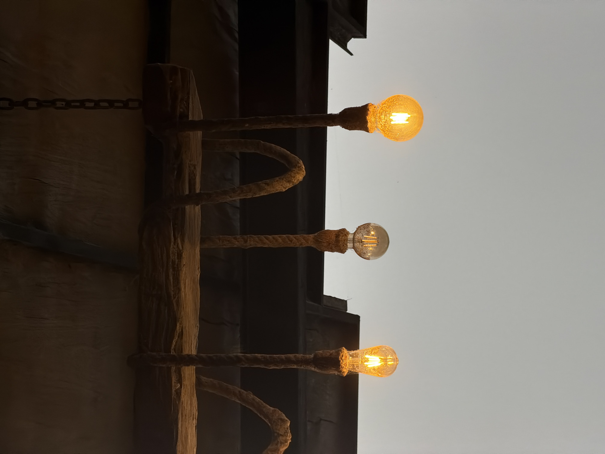 A close-up, low-angle shot shows three incandescent light bulbs hanging from a rough-hewn wooden beam by thick, textured ropes. The bulbs emit a warm, yellow glow. The background is a muted, overcast sky. The overall aesthetic suggests an outdoor or rustic setting, possibly a cafe or open-air market in Kintamani, Indonesia, given the lighting and materials. The image is primarily focused on the lighting fixture and its immediate surroundings, with no people or discernible actions visible.