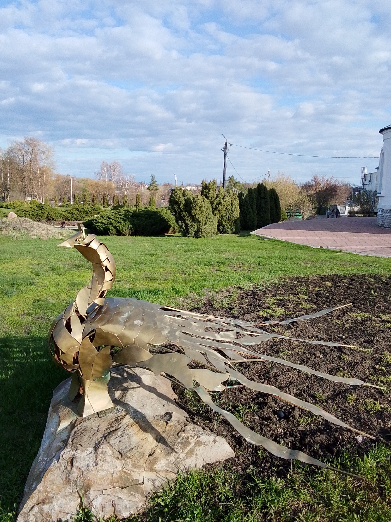 The image displays an outdoor scene featuring a prominent golden metal sculpture of a stylized bird, resembling a peacock or firebird, in the foreground. The sculpture is crafted from numerous individual golden-colored metal plates, forming its head, long neck, rounded body, and elongated, flowing tail feathers. It is positioned on a large, textured natural rock, with its tail feathers extending into a patch of dark, tilled earth.
The sculpture is surrounded by patches of bright green grass and garden beds. In the mid-ground, manicured evergreen bushes and hedges define parts of the landscape. Beyond these, a variety of deciduous trees, some still bare and others showing early signs of spring foliage, are visible under a bright, cloudy sky. A paved pathway, made of reddish-brown bricks, curves along the right side of the frame, leading towards a white building with a distinctive rounded turret that is partially visible. In the distance, two small figures, likely people, can be seen walking on the paved path. A utility pole with power lines is also present in the background.
The scene depicts a peaceful public park or garden in Khlevnoye, Russia, on what appears to be a clear, bright day, likely in spring or early summer given the emerging foliage and shadows cast by the sculpture. There is no visible text in the image.