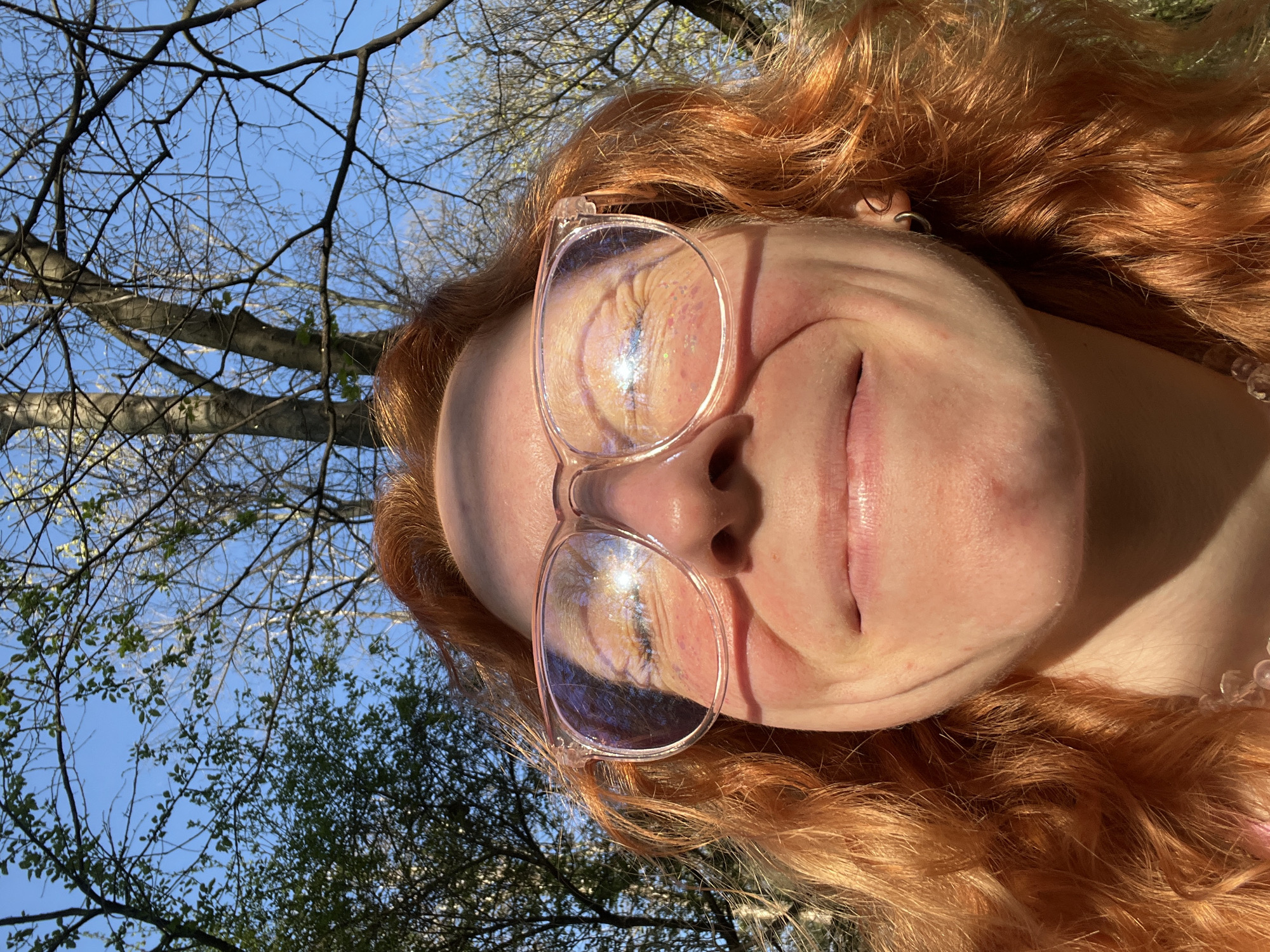 A person with red curly hair is pictured from a low angle, looking up at the sky. They are wearing clear-framed glasses that reflect the sky and trees. The background consists of tree branches against a blue sky. The location cannot be determined with the provided information.