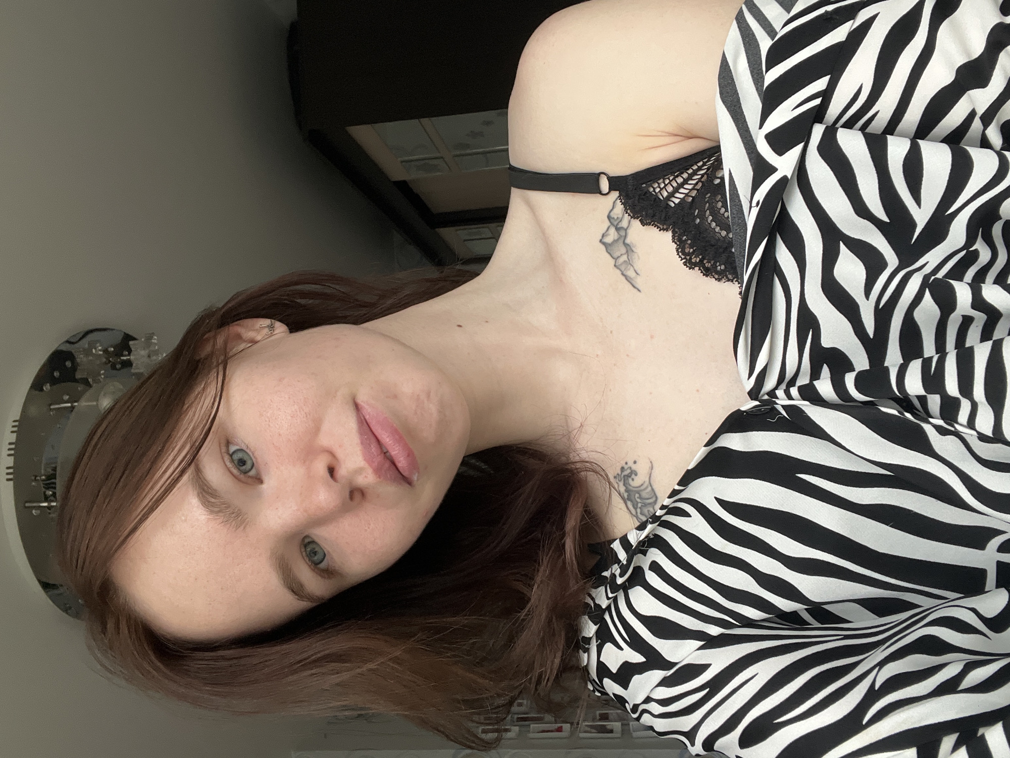 A woman is captured in a portrait-style photograph. She has fair skin, light blue eyes, and brown hair that falls around her face. She's wearing a black lace bra, visible around her shoulder and chest, and a zebra-striped garment covers part of her torso. A tattoo is visible on her chest, and another is on her collarbone. The background is softly lit and appears to be an interior space. To the left of the frame, a decorative clock or object with metallic elements is partially visible. The setting's location in the city of Chaikovsky, Russia cannot be determined from the image.