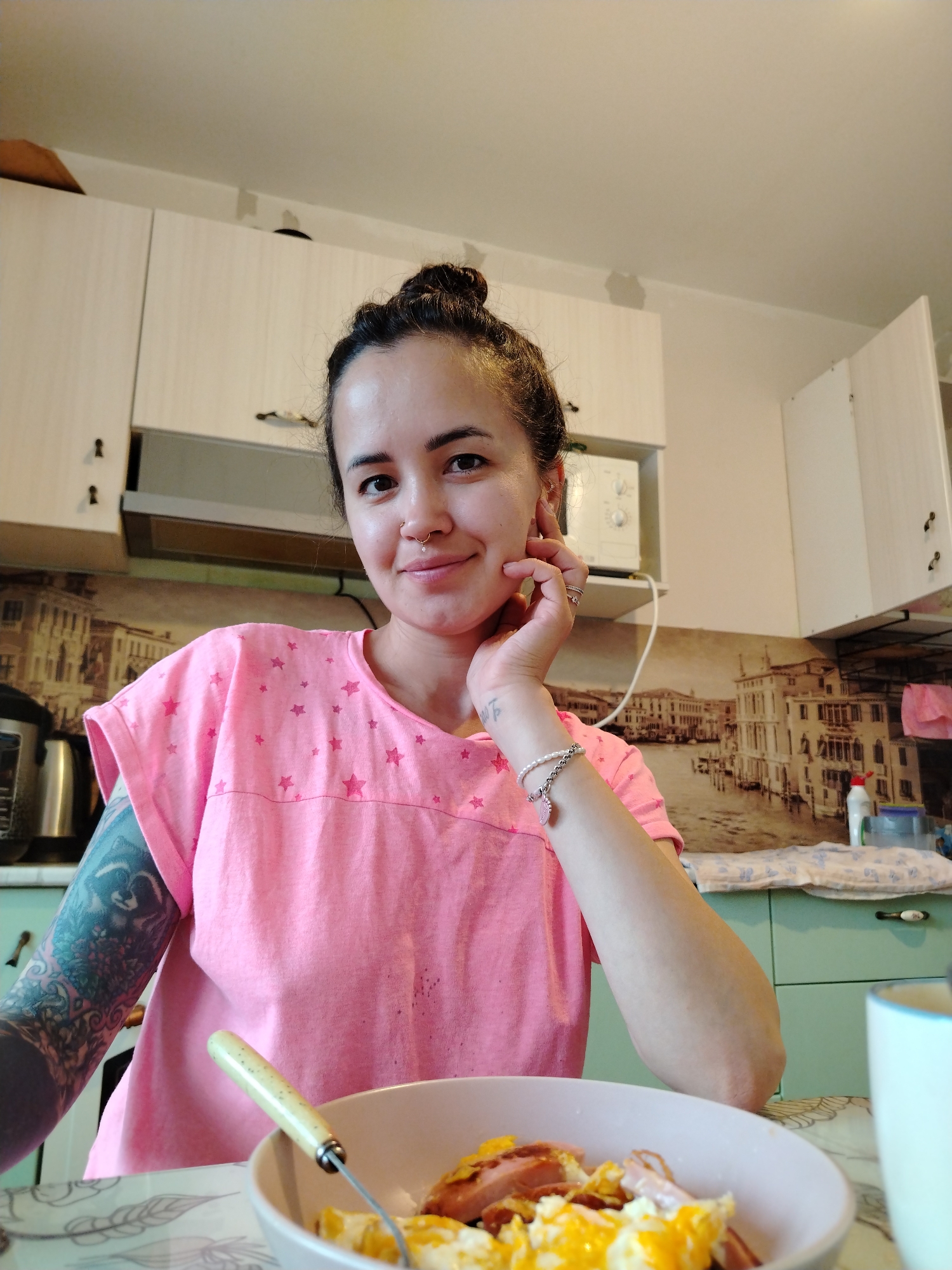 An adult female is seated at a table in a kitchen, identified as being in Noviy, Russia, looking directly at the camera with a slight smile. Her dark hair is pulled into a messy bun. She has a small, gold-colored hoop nose ring with a clear gem in her left nostril. Her left arm, featuring a detailed, dark blue/grey full-sleeve tattoo with a prominent raccoon motif on her upper arm, rests on the table. Her right hand, adorned with multiple rings on the ring finger and a bracelet appearing to be white beads and a silver chain with a heart charm, touches her chin/cheek. She wears a short-sleeved pink t-shirt with scattered darker pink star patterns.
In front of her, on a light-colored table with a subtle leaf/feather pattern, is a light-colored bowl containing a meal of scrambled eggs and pieces of sausage or ham, with a light-colored fork inserted. To her immediate right, a portion of a white and light blue mug is visible and blurred.
The background reveals a kitchen environment. White or light cream-colored upper cabinets with dark handles are mounted above a stainless steel range hood. To the right of the range hood, a white microwave rests on a shelf. A sepia-toned backsplash featuring a cityscape with canals and buildings, reminiscent of Venice, runs beneath the upper cabinets. The lower cabinets on the right are mint green or light teal, with a dark handle. On the right counter, a patterned fabric or mat is visible, along with a red-capped bottle and small containers. Above the cabinets, the wall is light beige/off-white, showing some faint, greyish smudges near the white ceiling. A silver electric kettle or similar appliance is partially visible on the left counter.