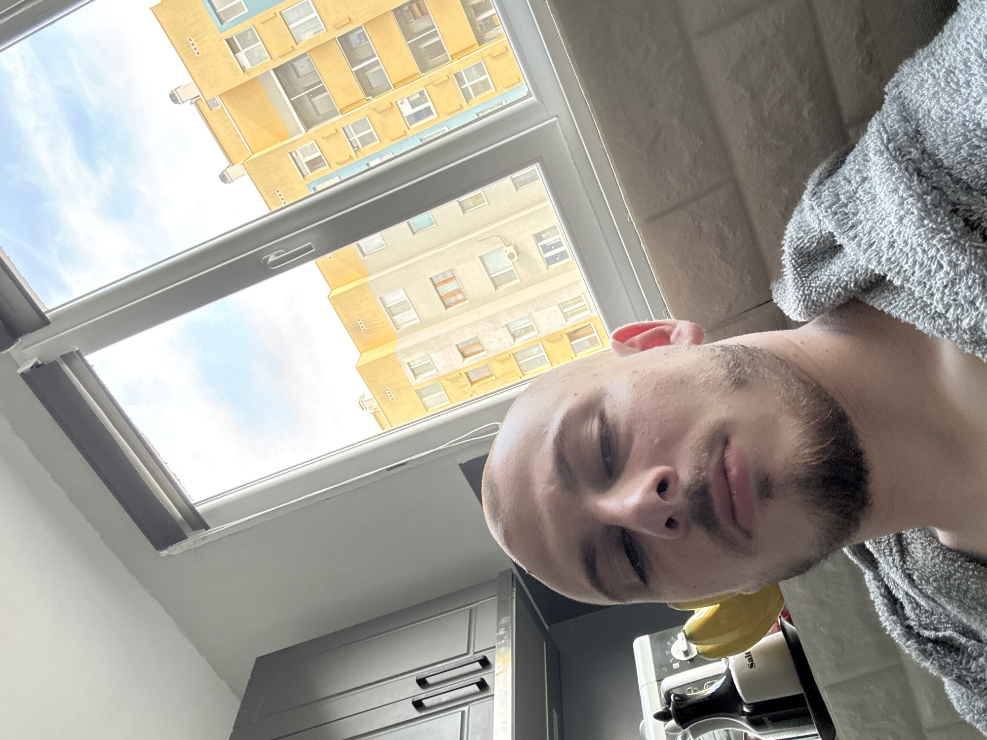 A selfie captures a young man, likely in his 20s or 30s, with a shaved head and light stubble beard, looking towards the camera with a neutral expression. He is wearing a grey, textured garment, possibly a bathrobe or towel. The image is taken from a low, rotated angle, giving it an unconventional perspective.
The scene appears to be an indoor space, likely a kitchen, featuring a large, white-framed skylight or window. Through this window, a bright blue sky with scattered clouds is visible, indicating daytime. Beyond the sky, several multi-story residential buildings are seen, characteristic of an urban environment. These buildings are predominantly yellow or ochre in color, with some featuring light blue accents around windows or balconies, consistent with architecture found in Stara Zagora, Bulgaria.
Within the indoor space, dark grey kitchen cabinets with horizontal handles are partially visible on the left side of the frame, below the window. On the right, a kitchen counter holds a light-colored appliance, possibly a blender or coffee machine. Next to this appliance are several yellow bananas and a white shaker clearly labeled "Salt". A small pink fabric item is also partially visible near the man's shoulder. The bright illumination suggests clear weather during the day, possibly morning or early afternoon.