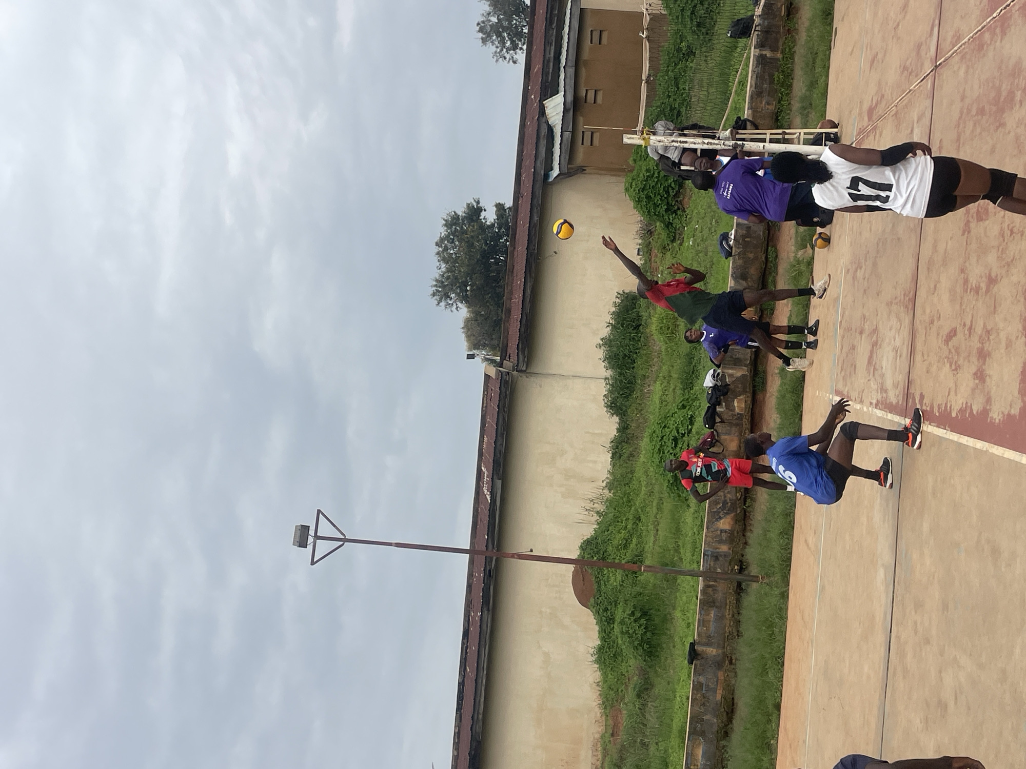 In Gura Top, Nigeria, a volleyball game is in progress on an outdoor court under an overcast sky. A group of individuals, predominantly young adults, are engaged in the sport. One male player, wearing a red and green jersey, is in mid-air, attempting a spike with a yellow and blue volleyball. Several other players are on the court, some in defensive stances and others observing the play. The court is made of concrete, with distinct lines marking boundaries. In the background, a weathered, light-colored building with a corrugated metal roof stands behind a low concrete wall and overgrown green vegetation. A tall, rusty metal pole with a light fixture is positioned to the left of the frame. The scene depicts a casual athletic activity in an outdoor urban setting.