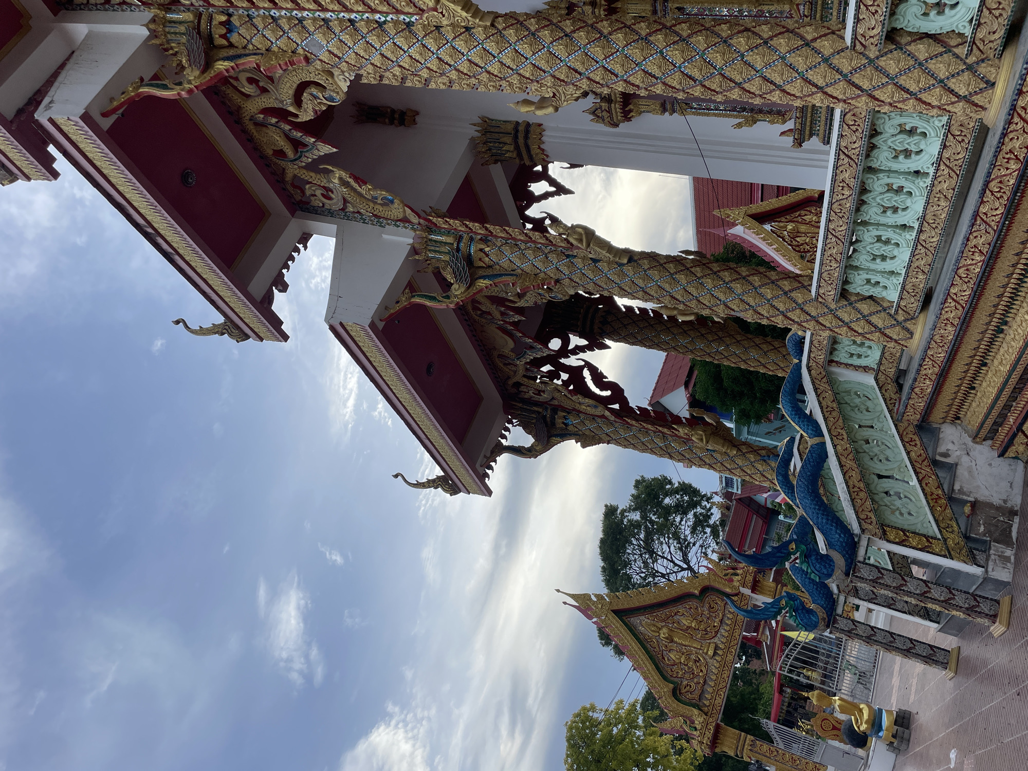 The image shows the exterior of a Buddhist temple, likely located in Pattaya City, Thailand. The temple features ornate architecture with vibrant colors and intricate details. Golden pillars adorned with mosaic tiles and sculpted figures rise towards a red and gold roofline. A staircase with intricately designed railings, featuring blue serpent-like statues (Nagas) at the top, leads up to the temple entrance. In the background, other temple structures and trees are visible under a cloudy sky. The overall scene suggests a place of religious and cultural significance.