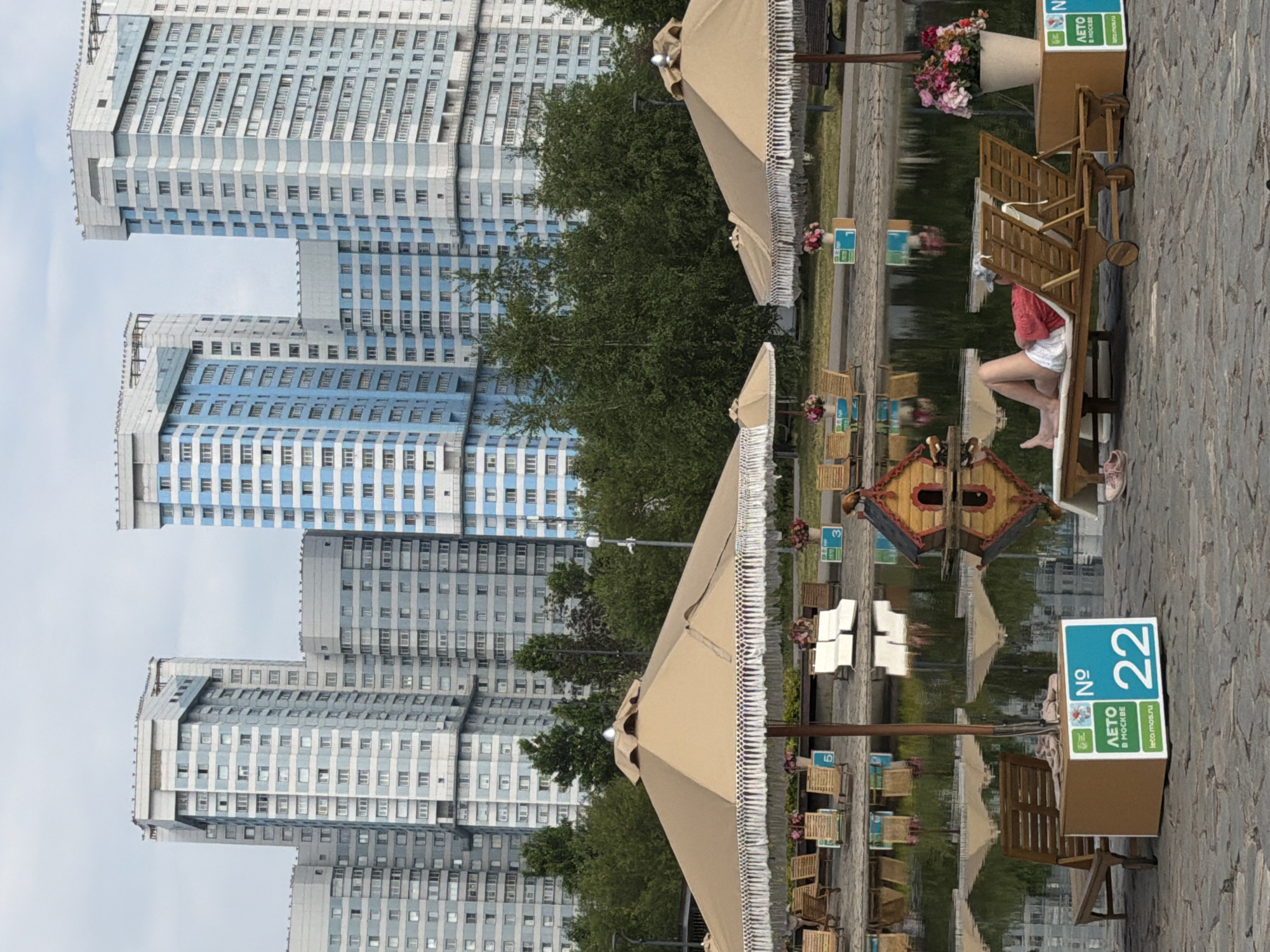This image depicts an outdoor relaxation area with a body of water in the foreground and a complex of residential buildings in the background, likely located in Khimki, Russia. Several large, tan umbrellas with decorative fringe are set up over a paved area. In front of the umbrellas are rows of wooden lounge chairs. A person is lying on one of these chairs, facing away from the camera. The water reflects the umbrellas, chairs, and buildings. There are also some small decorative elements, including a miniature wooden house that resembles a birdhouse and pots of flowers. In the mid-ground, two blue and white signs are visible, one with the number "22" and the other with the number "3". The buildings in the background are tall, multi-story residential blocks with a distinct architectural style featuring blue and white vertical stripes. The sky is overcast.