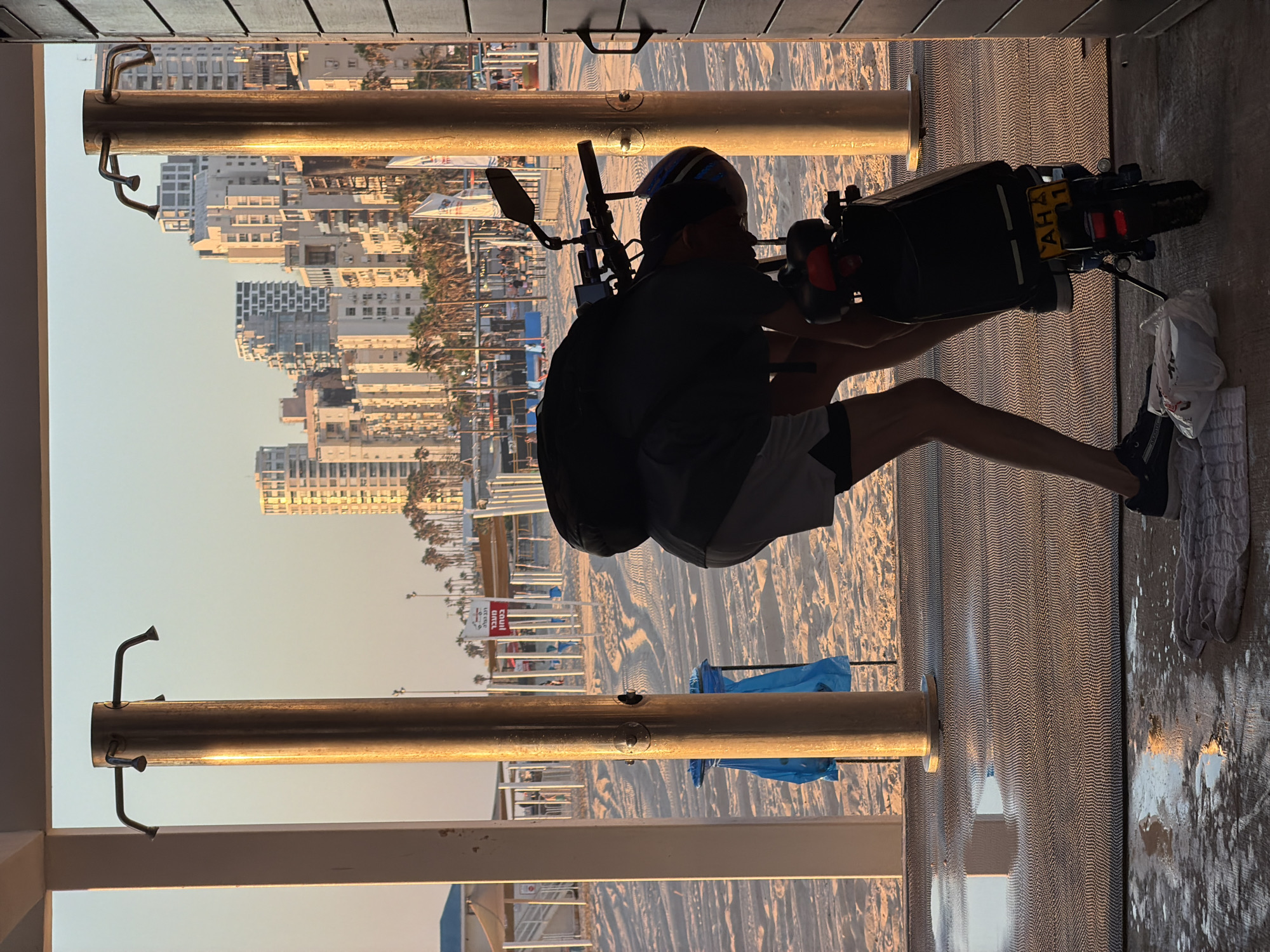 The image depicts a scene on a beach in Bat Yam, Israel, at sunset. In the foreground, a person is bent over an electric scooter, possibly making adjustments or preparing it for use. The person is wearing shorts, a t-shirt, a backpack, and a helmet. The scooter has a license plate with the letters "AHA" and the number "01." A towel and a plastic bag lie on the wet, patterned ground in front of the scooter.
Behind the person and the scooter, a large metal pole with showerheads is visible, suggesting the presence of beach showers. In the background, the sandy beach stretches out, marked with tire tracks. Beyond the beach, a line of palm trees and modern high-rise buildings forms the city skyline. The sky is a warm, golden hue, indicative of the setting sun. The overall impression is one of a tranquil beachside setting at the end of the day, with urban elements visible in the distance.