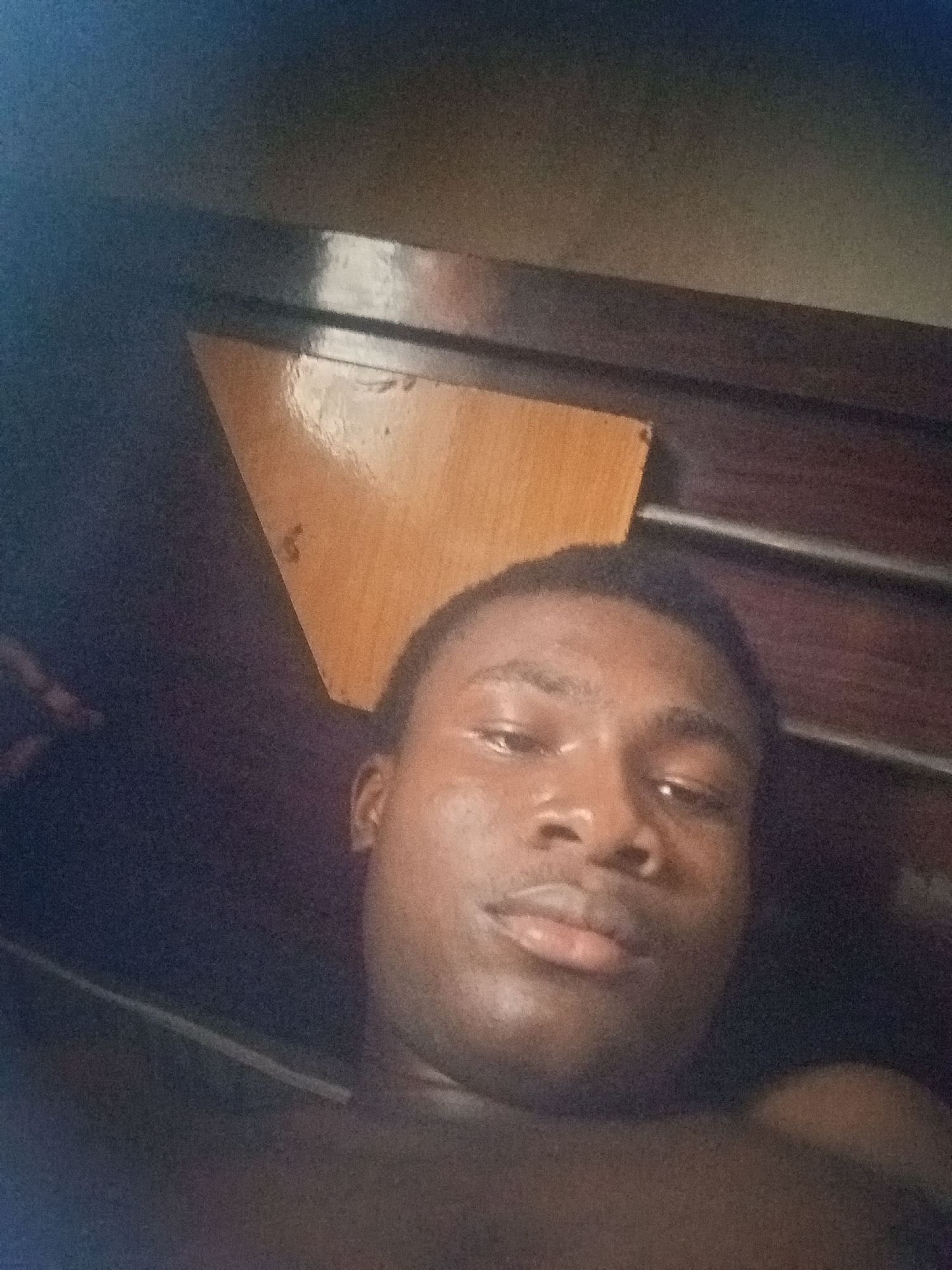 An amateur, close-up selfie of a young Black male is taken from a low angle. The man is lying down, his head tilted slightly back, and he is looking towards the camera with his eyes open. The background consists of a wooden surface with a triangular section of lighter wood in the upper left quadrant, and a dark, textured surface that appears to be a wall or furniture. The lighting is dim, creating soft shadows on the man's face. The image is grainy, indicating it was taken in low light conditions with a low-quality camera. Based on visual cues, this image was likely taken indoors, in a residential setting in Nkpor, Nigeria.