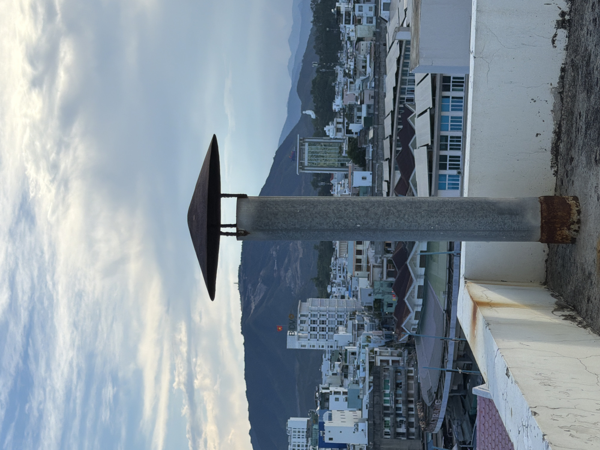 The image is a landscape photograph taken from a rooftop in Nha Trang Ward, Vietnam, looking out over a city with hills in the background. In the foreground, a tall, weathered metal pipe with a conical cap stands prominently. The pipe shows signs of rust near its base. The rooftop itself is made of concrete and has a white, cracked wall along its edge, with some staining and peeling paint. Beyond the rooftop, the cityscape unfolds with numerous buildings of varying heights and architectural styles, many with light-colored facades and flat or tiled roofs. Some buildings are modern, with glass windows, while others appear older. A prominent white building with the "BIDV" logo and a Vietnamese flag is visible in the mid-ground. The background features rolling, dark green hills under a cloudy sky. The overall lighting suggests it is either early morning or late afternoon. There are no people or animals visible in the image.