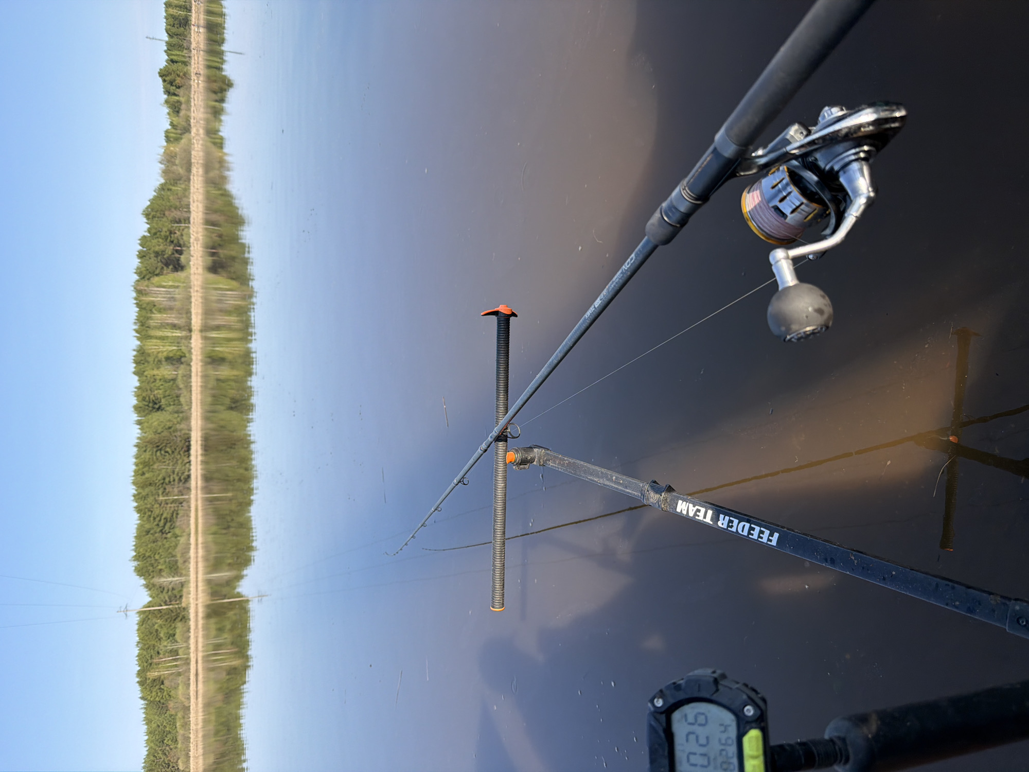 The image shows a fishing rod and reel set up on a bank in Chaikovsky, Russia. The rod is held in place by a rod rest with an orange top. A line from the fishing rod goes into the water. Reflected in the water is the opposite bank with trees and a clear blue sky above. The words "FEEDER TEAM" are visible on the rod rest. A digital device is partially visible in the lower left, showing numbers.