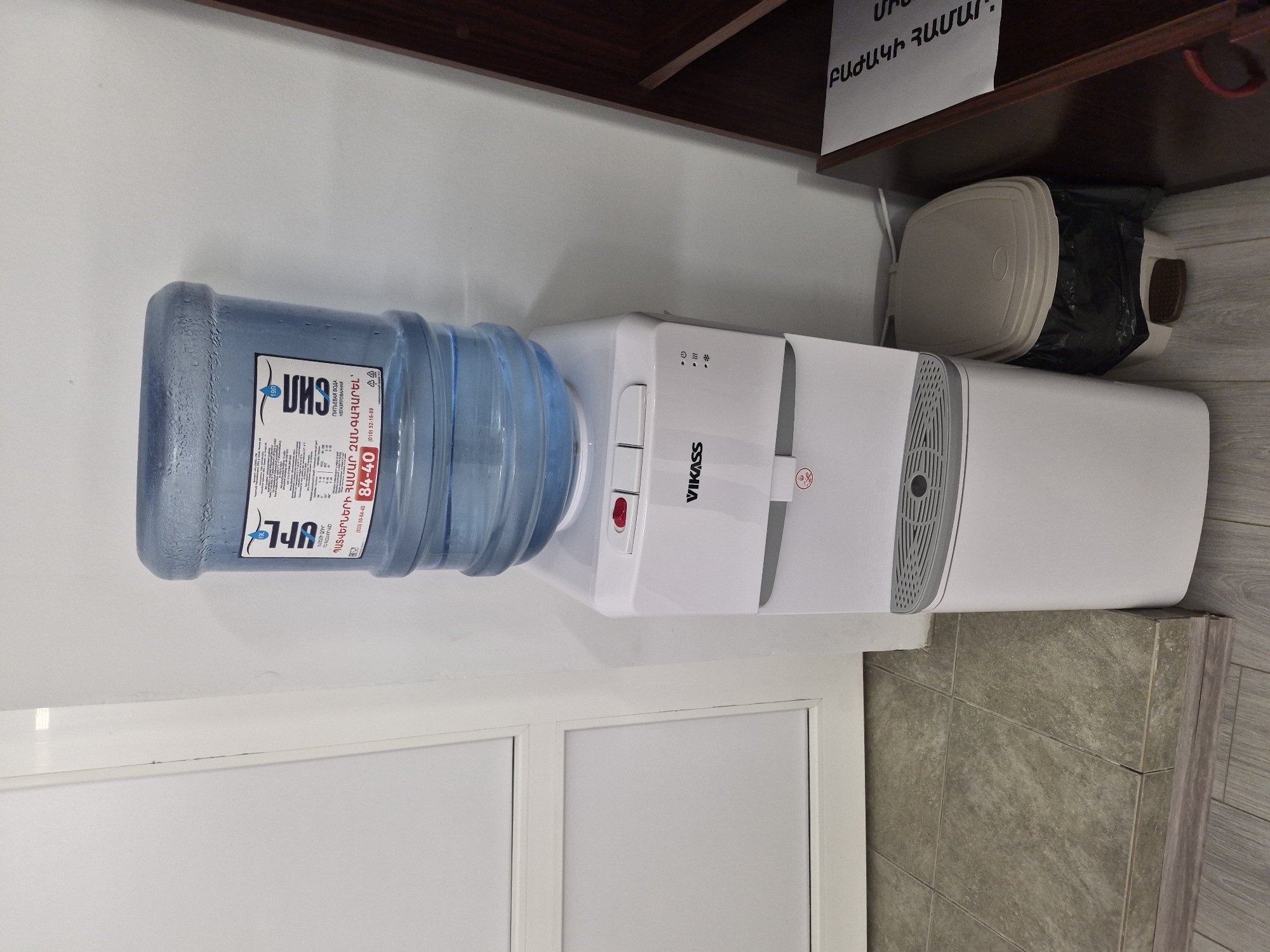 An indoor scene showcases a white water dispenser with a large, inverted blue plastic water bottle, positioned against a white wall. Above the dispenser, a dark brown wooden shelf is visible, with a small, beige-colored trash bin tucked underneath, containing a black plastic bag. A piece of white paper with Armenian text is taped to the underside of the shelf.
The water bottle's label displays Armenian text: "ՍՈՒՐԲ" (Surb), "ԵԼԵԼՈՒ ՋՈՒՐ" (Drinking water), and "ՊԱՏՎԵՐՆԵՐԻ ՀԱՄԱՐ ԶԱՆԳԱՀԱՐԵԼ՝" (To order, call:), along with phone numbers (093) 00-84-40 and (010) 52-16-09. It also indicates "19լ" (19L). Russian text "ПИТЬЕВАЯ ВОДА НЕГАЗИРОВАННАЯ" (Drinking water, non-carbonated) is present. The water dispenser is branded "VIKASS" and features a prominent red dispensing button, along with indicator lights for power, heating, and cooling. The paper on the shelf reads "ԲԱԺԱԿԻ ՀԱՄԱՐ," (For the cup).
The background includes a white door frame on the left and a gray tiled floor or wall section on the right. The scene is static, devoid of people or direct activity, suggesting a functional area such as an office or public space in Yerevan, Armenia.