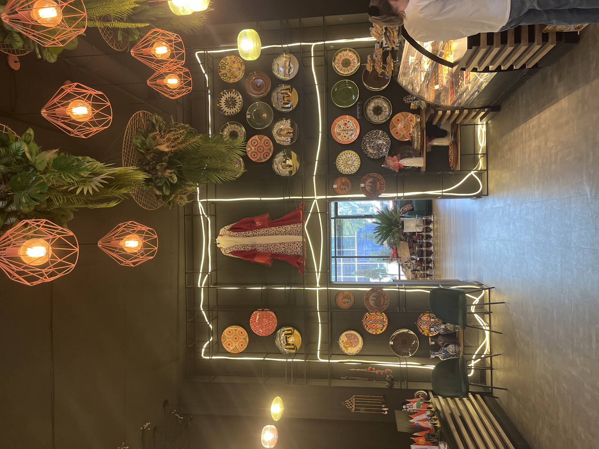 The image, rotated 90 degrees counter-clockwise, presents the interior of a commercial establishment, likely a cafe or shop, characterized by its dark walls and warm, eclectic decor.
On the left side of the frame, several light fixtures hang from the ceiling. Some are bare bulbs in geometric, coral-colored wire cages, while others are simple glass pendants. Artificial green plants and foliage are arranged in hanging baskets and wall-mounted planters, adding a natural element.
The central feature is a large dark wall adorned with a collection of decorative plates of various sizes, patterns, and colors, mounted on a grid structure. Winding neon-like LED strips highlight sections of this display. A prominent red and white embroidered garment, possibly a traditional caftan, is dramatically hung on this wall.
To the right, a person wearing a white t-shirt and blue jeans stands behind a counter with a wooden slat facade and a glass display case filled with food items, likely pastries or desserts. The light-colored floor, possibly concrete, stretches towards the foreground where two dark green upholstered chairs are visible, suggesting a seating area.
In the background, a large window provides a view of an exterior building with blue glass and daylight, indicating the time of day. Small flags of various countries are displayed on a shelf in the lower right corner, alongside other decorative objects. No legible text is visible in the image. The overall ambiance is cozy and inviting, with a blend of modern lighting and traditional decorative elements.