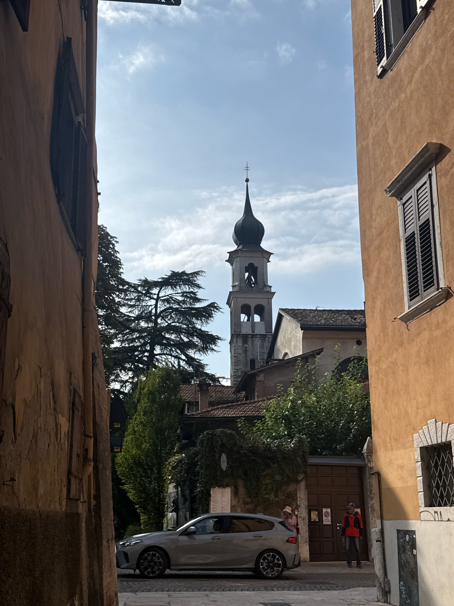 The image shows a view down a narrow street in Trento, Italy. On the right, a tall, orange-colored building with closed shutters on its windows frames the scene. On the left, a building with weathered, textured walls partially obscures the view. In the background, a church tower with a dark, pointed spire rises above other buildings and trees. A silver sedan is parked on the cobblestone street. Two individuals are visible: one wearing a hat and a pink top stands near the car, and another person in a red jacket and blue jeans stands near a large wooden door. Lush green trees and foliage add to the urban landscape. The sky is blue with scattered white clouds.