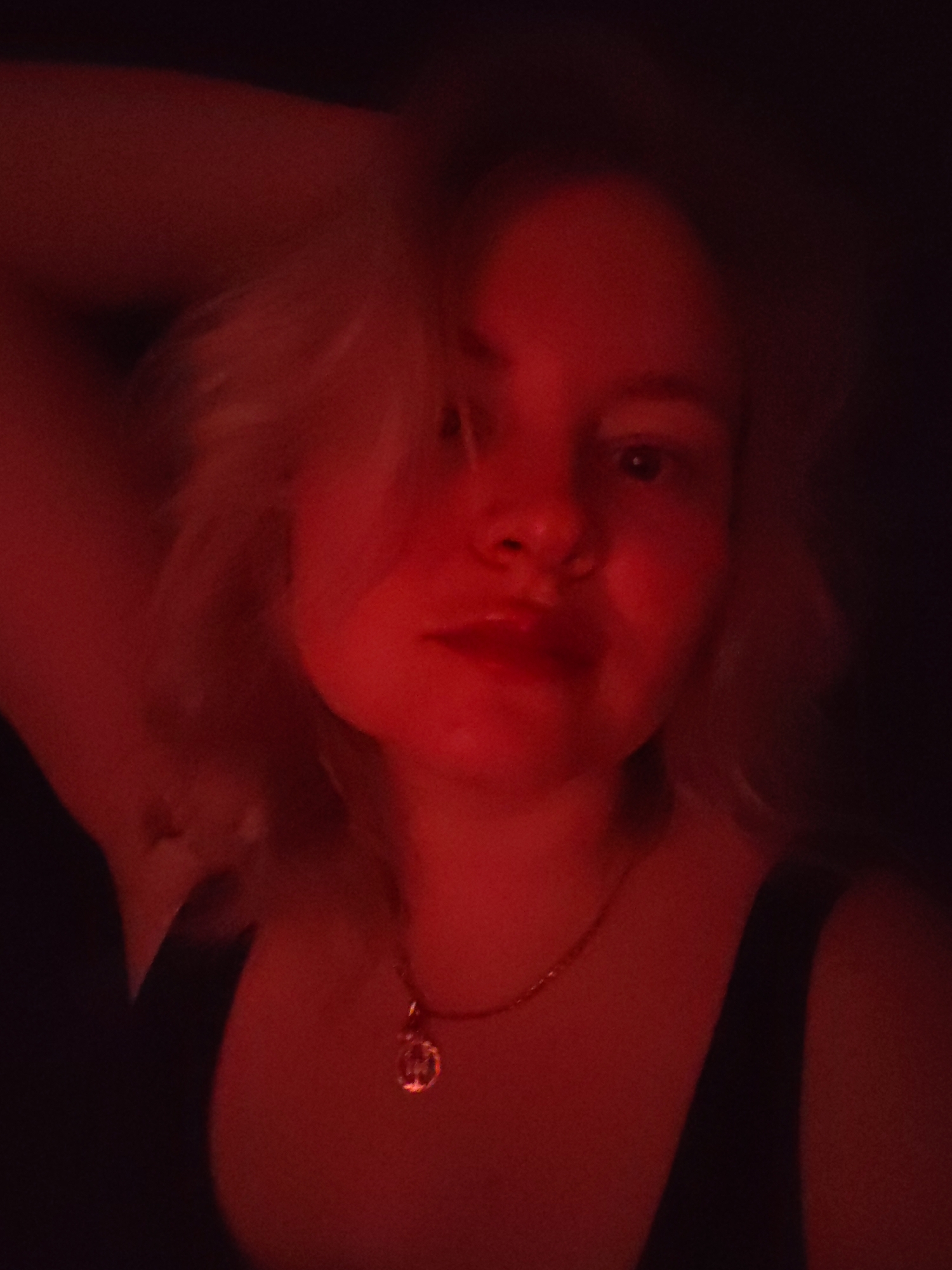The media file presents a dimly lit, close-up portrait of a young woman, likely a selfie. The entire image is bathed in an intense red light, which creates a dramatic, warm, and somewhat mysterious atmosphere against a dark, featureless background. This strong artificial lighting suggests an indoor setting, likely at night.
The central subject is a woman, appearing to be in her 20s or early 30s, with light-colored hair, possibly blonde or light brown, which is styled with volume around her face. Her facial features are softly illuminated by the red glow; her eyes appear light, and her lips are slightly parted, conveying a neutral or gently contemplative expression.
She is dressed in a dark, likely black, sleeveless top or tank top. Around her neck, she wears a delicate gold chain necklace adorned with a small, round or coin-shaped pendant. Her left arm is visible, raised with her hand positioned behind her head, suggesting a posed shot. There are no discernible details in the background, only deep shadows. No text is visible within the image. The context provided indicates the location is Izhevsk, Russia.