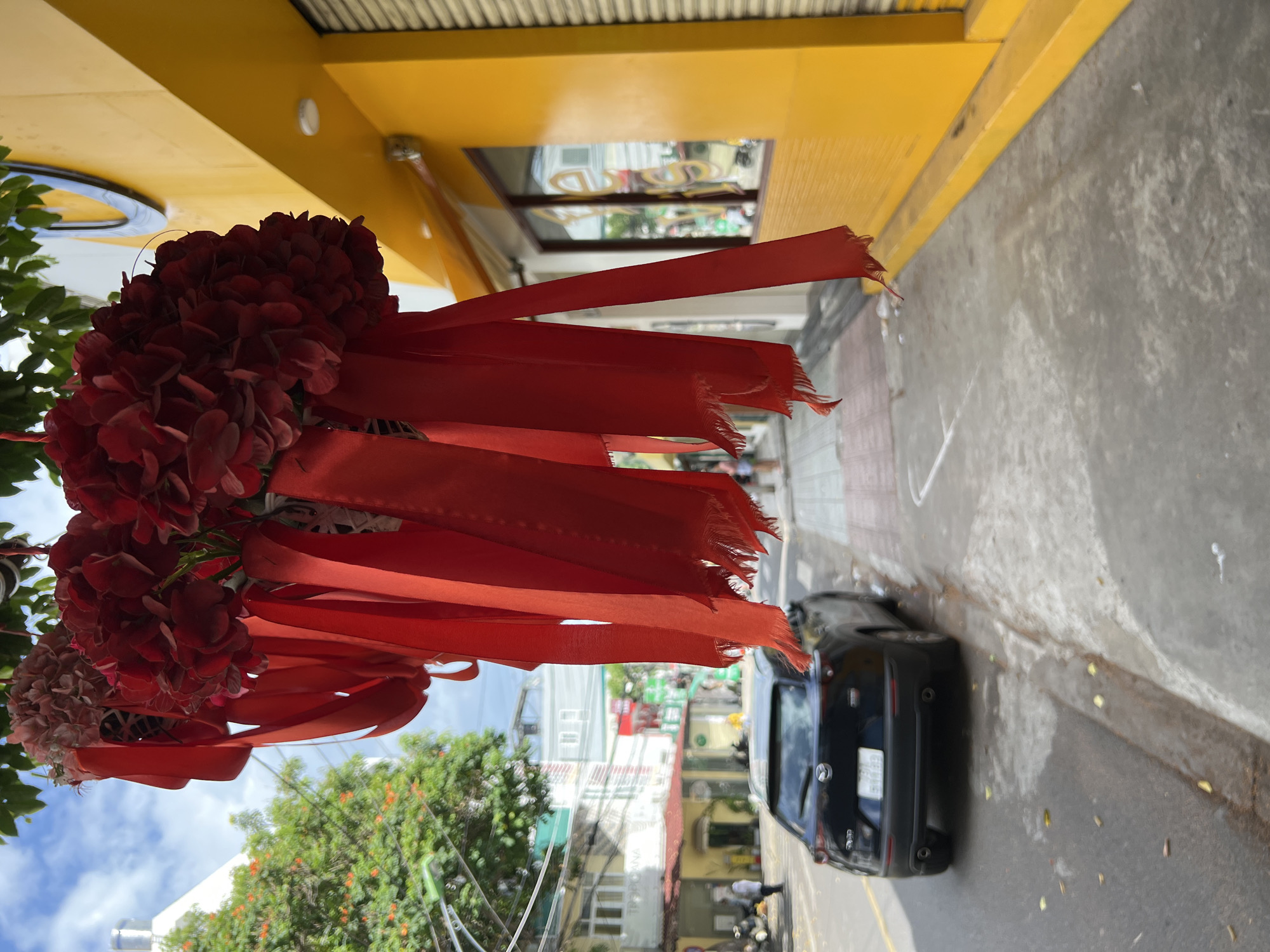 In Nha Trang, Vietnam, on a street in front of a bright yellow building with large windows, a black Mazda CX-30 is parked near the curb. The car's license plate is visible, reading "51G-810.88". The street is paved, and a sidewalk runs alongside the building. Hanging from an unseen overhead structure, a cluster of artificial red flowers adorned with red ribbons partially obscures the view of the building and the street. The ribbons, frayed at the ends, hang loosely, some touching the sidewalk. Green trees line the street in the background, and a cloudy blue sky is visible above. The building's facade is yellow, with horizontal blinds covering part of the windows.