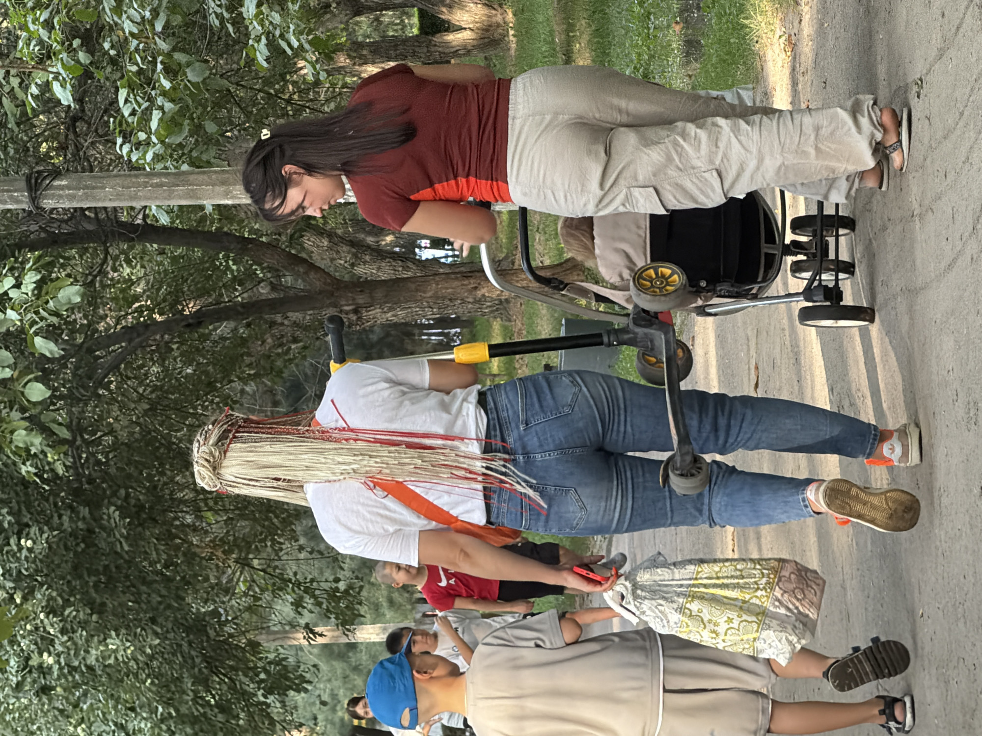 In a park-like setting, likely on a paved path, a group of people are walking. In the foreground, a child wearing a blue cap and a grey shirt and shorts walks to the left, carrying a patterned bag in their right hand. To their right, a woman with long blonde braids walks away from the camera, wearing a white t-shirt and blue jeans. She is holding a red object in her right hand. Next to her, a woman with dark hair is pushing a stroller containing a baby, and she is wearing a red and brown shirt and light-colored cargo pants. Behind the child in the blue cap, another child in a red shirt and dark shorts walks. Further back, other individuals can be partially seen. The environment consists of trees and foliage, suggesting an outdoor public space. A utility pole is visible on the right side of the image.