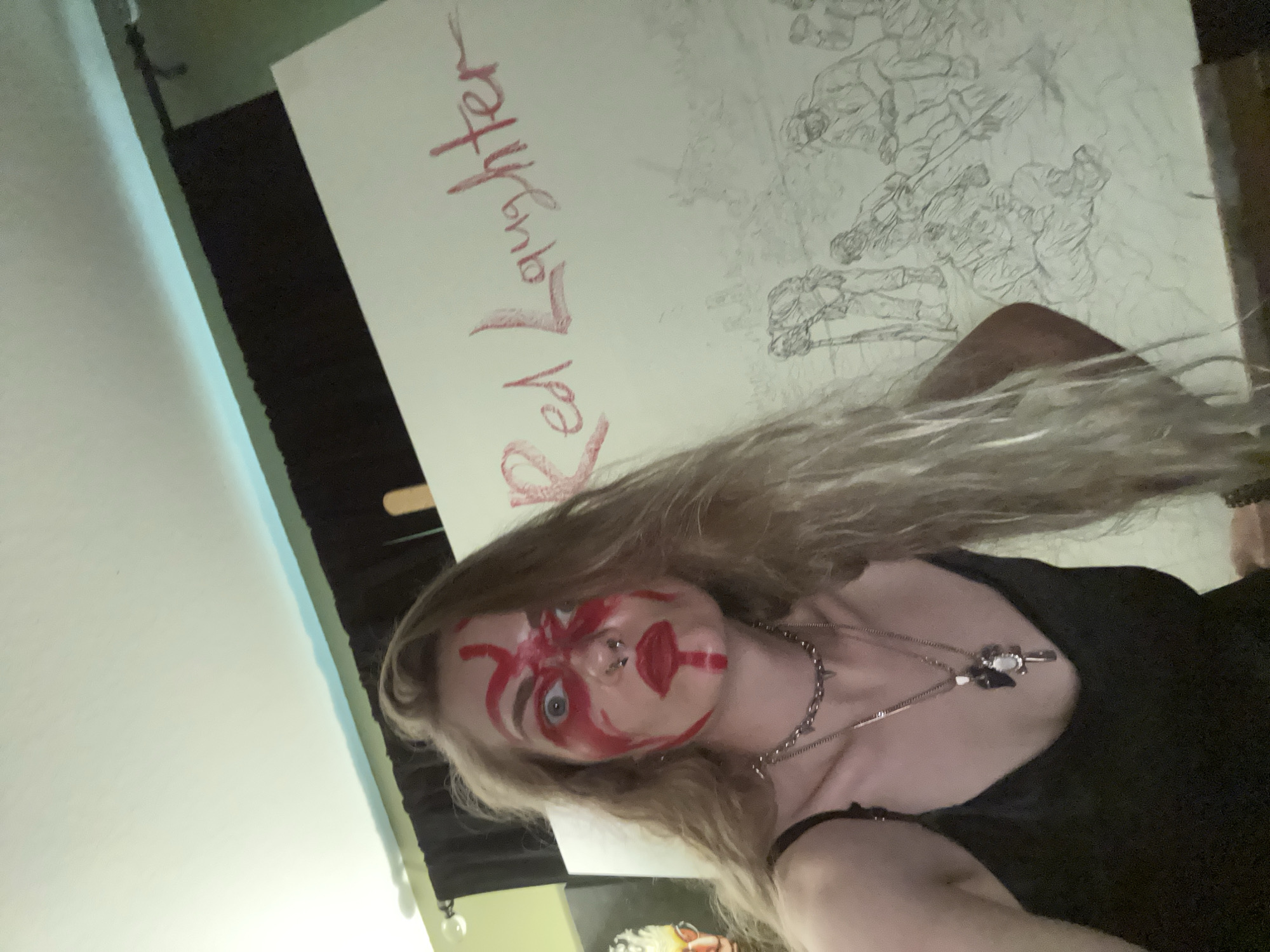 A woman with long blond hair and red paint on her face in a tribal-like pattern stands in front of a large canvas. The canvas displays a drawing of multiple figures and the words "Red Laughter" written in red. She is wearing a black top and multiple necklaces. The setting appears to be an indoor studio or living space. The location is the city of Cape Coral, United States of America, as requested by the user.