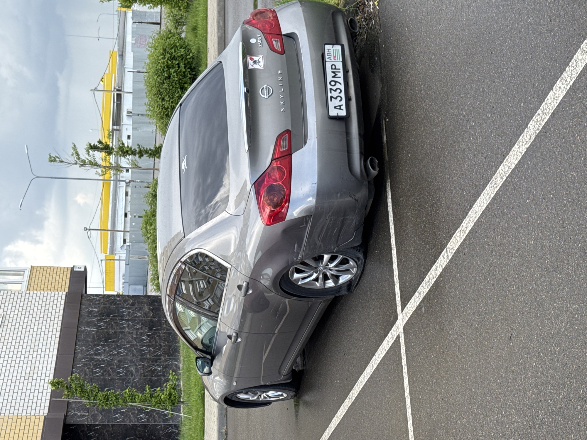 A gray Nissan Skyline sedan with a flat tire and damage to the rear bumper is parked in a parking space marked with white lines. The license plate reads "A339MP" with "ABH" indicating the region. Stickers are visible on the rear of the car. A building with brick and dark stone cladding is nearby, as is a commercial structure with yellow supports. The location is suggested to be Krasnodar, Russia.