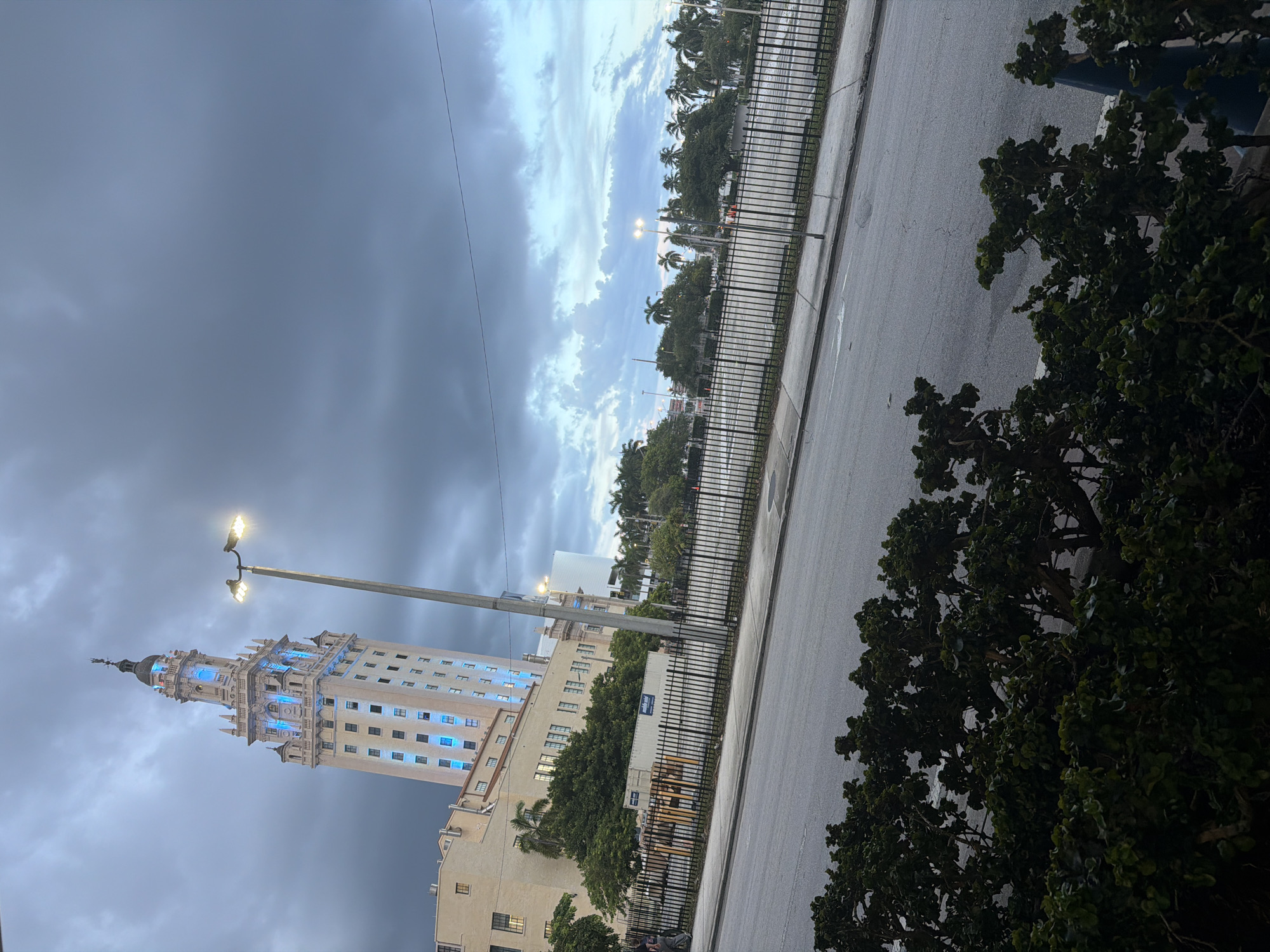 The image captures the Freedom Tower in downtown Miami, Florida, under a cloudy sky. The iconic tower, illuminated with blue lights, stands tall against the overcast backdrop. In the foreground, a street and a black metal fence enclose an open area with trees and palm trees visible beyond. A lamppost with bright lights stands to the right of the tower. The scene appears to be set in the late afternoon or early evening, given the dim lighting and the illuminated building.