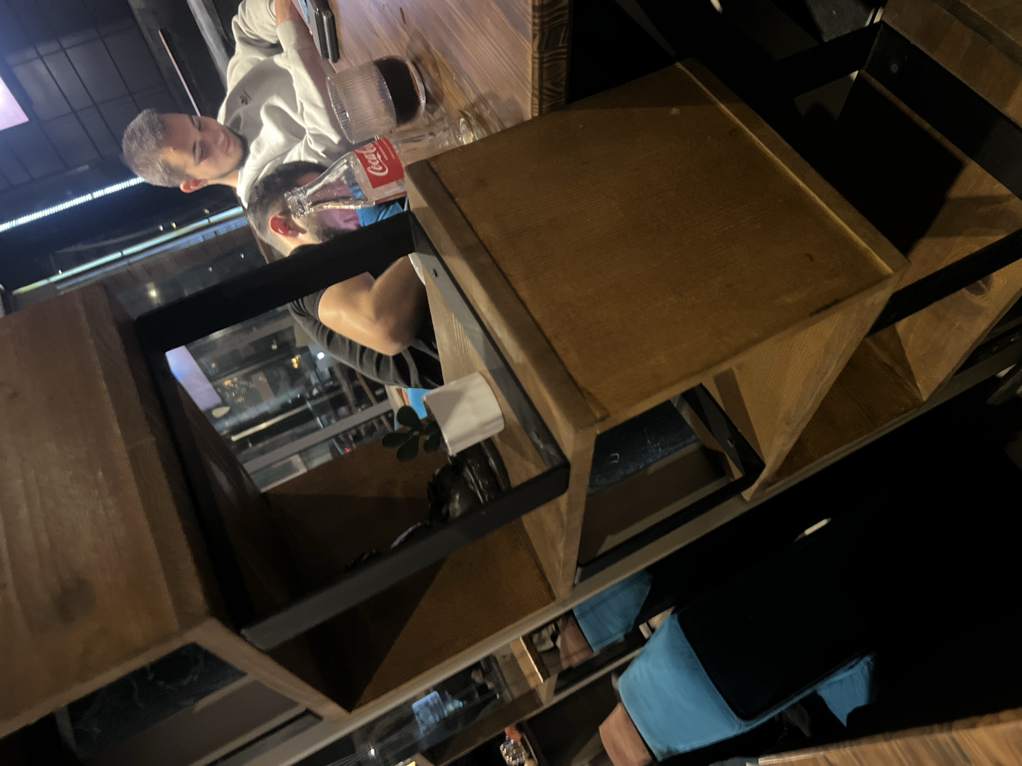 The image shows the interior of a restaurant or bar, likely located in Asenovgrad, Bulgaria, although this cannot be definitively determined from the image alone. Two men are visible at a wooden table. One man is wearing a light grey hoodie and is looking at a phone or tablet. The other man is wearing a dark shirt and appears to be holding a white napkin. On the table are a Coca-Cola bottle, a glass, and other items. The table is part of a wooden structure with shelves or compartments, and seating with black and blue cushions is visible. The environment suggests a modern or industrial aesthetic.