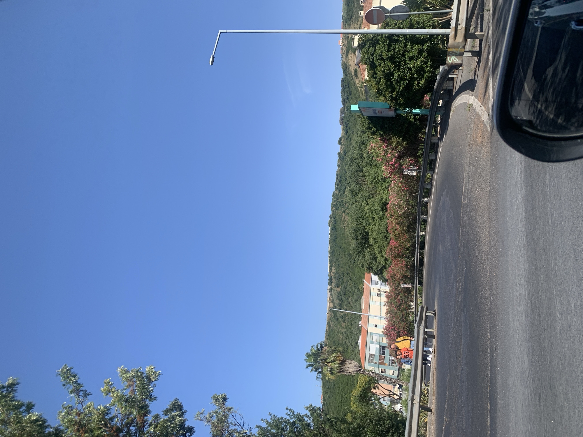 The image captures a sunny daytime scene along a curved road in Odivelas, Portugal, with a clear blue sky dominating the upper portion of the frame. In the foreground, the dark asphalt of the road is visible, with a reflective side mirror of a vehicle taking up the bottom right corner. A metal guardrail delineates the side of the road.
Further back, two individuals are walking on a paved area next to the road, near a palm tree. Behind them, a row of buildings with light-colored facades and orange tiled roofs lines the street. These buildings are nestled against a lush green hillside covered in trees and vegetation. Scattered amongst the greenery are patches of bright pink flowering bushes. A tall, modern street lamp stands prominently on the right side of the road, and a traffic sign with a red circle is visible further down. The environment suggests a suburban or semi-rural setting within Odivelas.